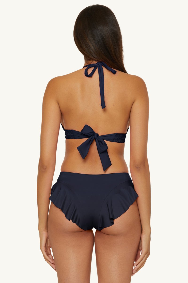 Woman with long brown hair is seen from behind, wearing an ink-colored halter bikini top and the Ruffle Hipster Swim Bottom - Monaco in Ink, featuring high-waisted ruffled details, against a plain background.