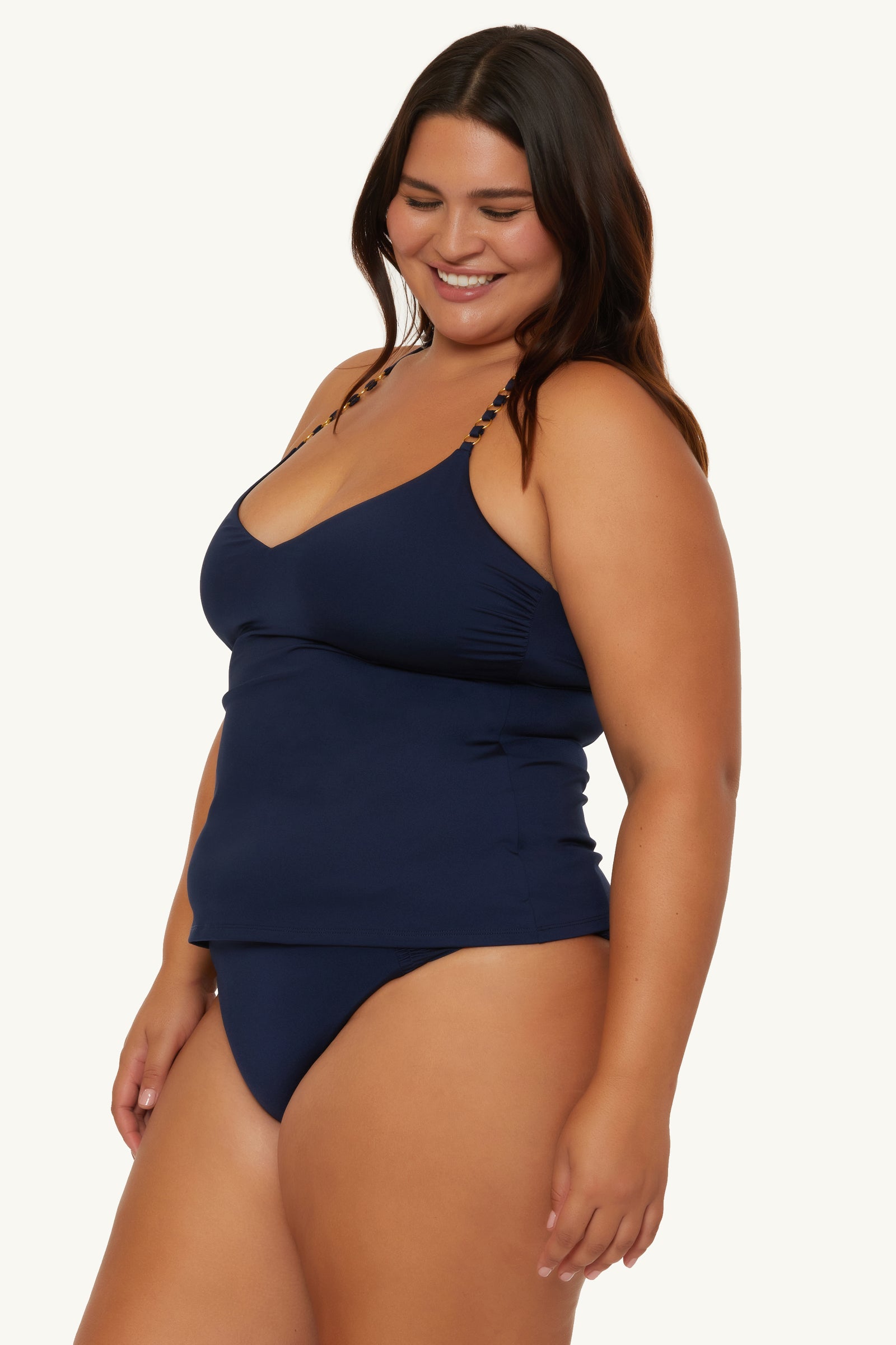 A woman with long dark hair smiles wearing the Ring Tankini Swim Top - Monaco, a navy blue swimsuit with thin straps and matching bottoms, standing against a light background.