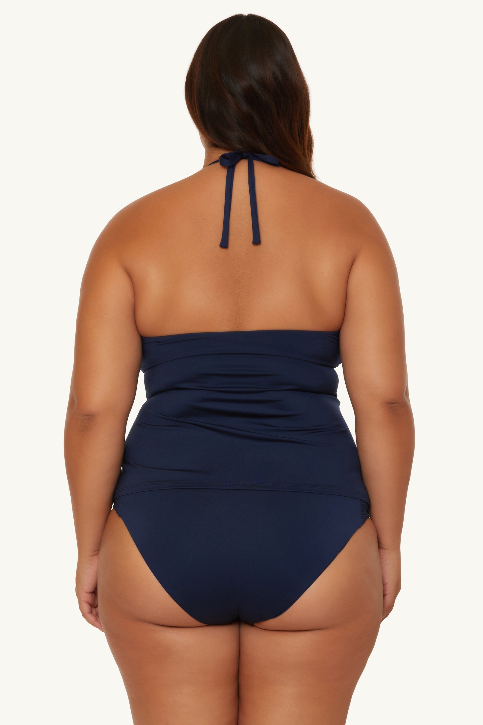 A woman with long dark hair is seen from the back, wearing the Ring Tankini Swim Top - Monaco in navy blue, featuring a halter-neck and ruched detailing against a plain, light background.