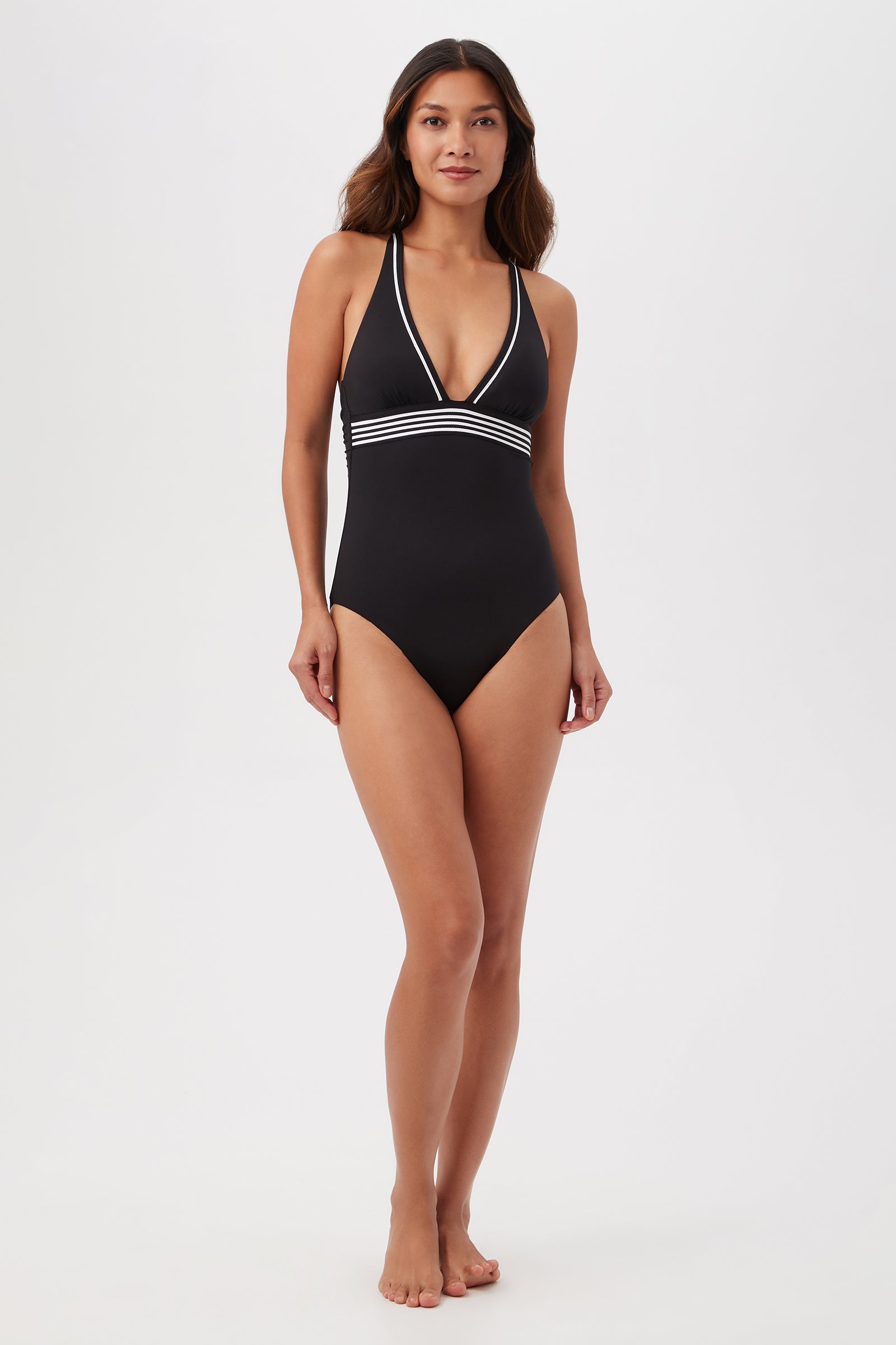 A woman stands barefoot against a plain white background, modeling the Monaco Pleated Plunge One-Piece—a black halter swimsuit with a deep V-neckline and white striped trim under the bust.