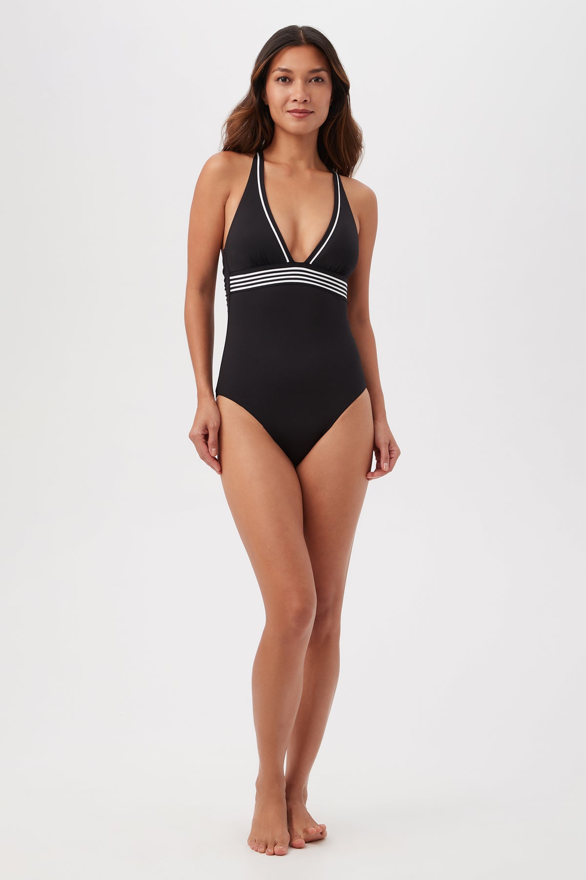 A woman stands barefoot against a plain white background, modeling the Monaco Pleated Plunge One-Piece—a black halter swimsuit with a deep V-neckline and white striped trim under the bust.