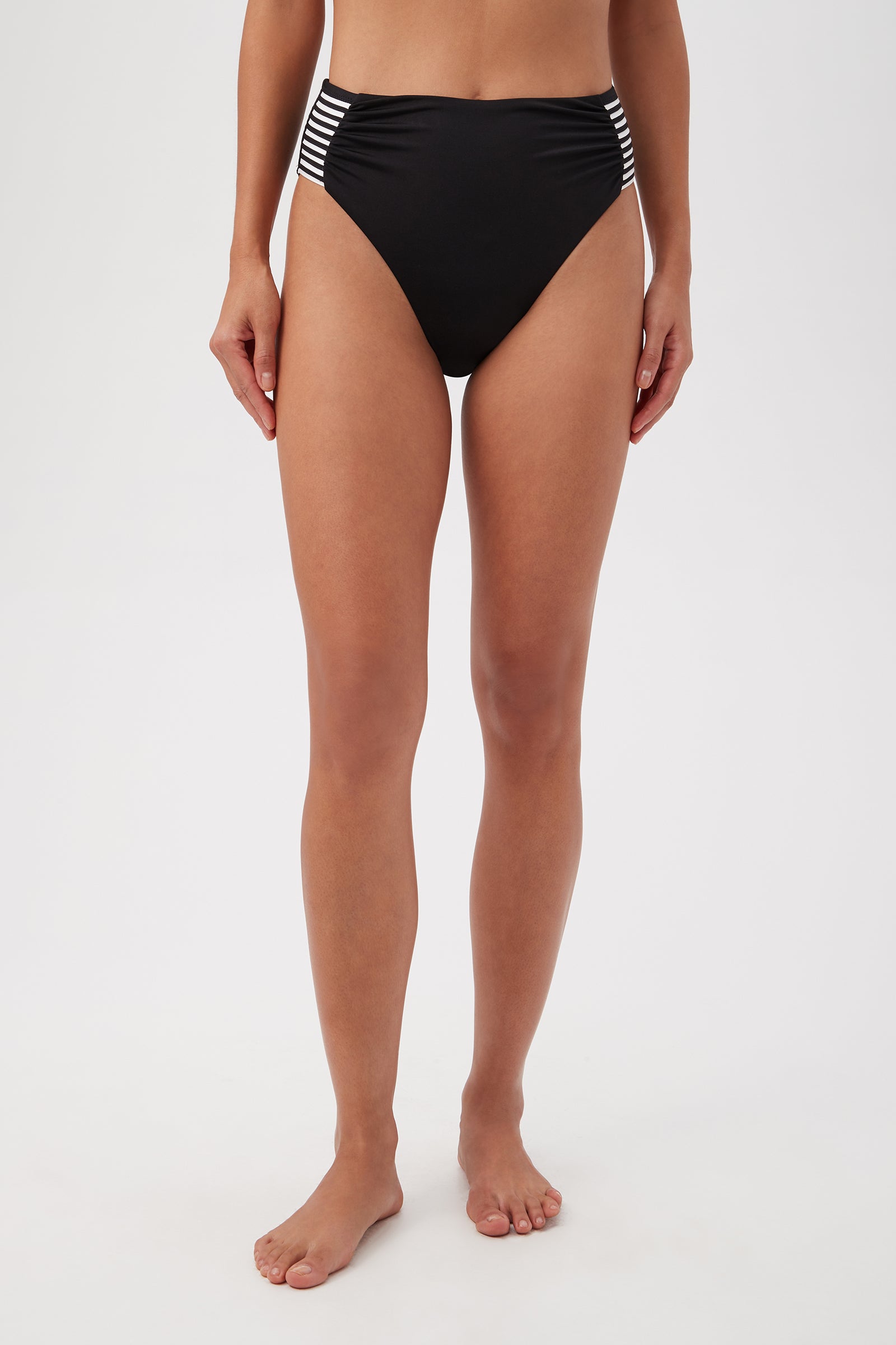 A woman models the Monaco Pleated High-Waist Bikini Bottom in Black/White, featuring vertical stripes on the sides, shown barefoot from midriff to feet against a plain white background.