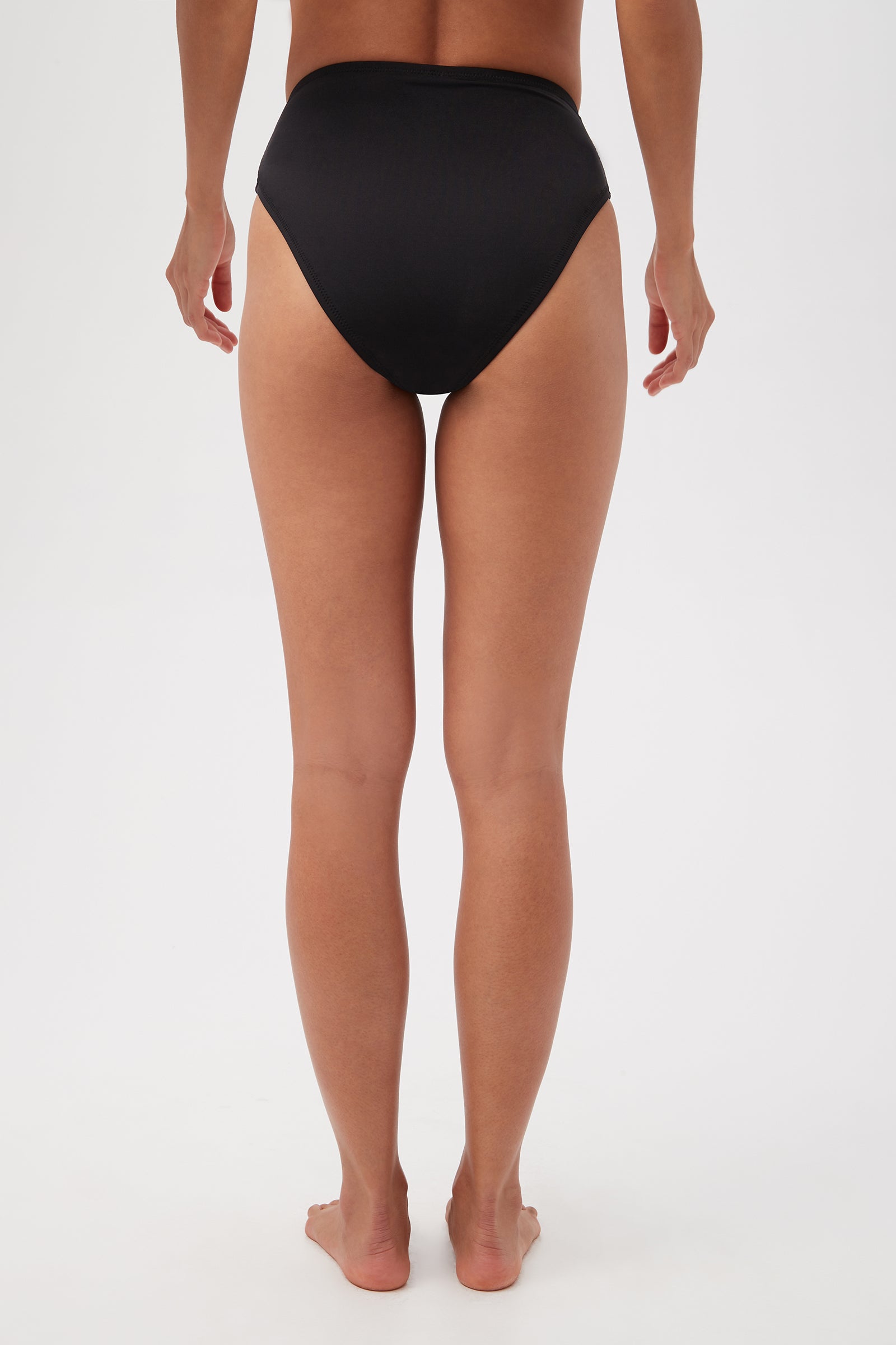 A person stands barefoot, facing away from the camera, wearing the Monaco Pleated High-Waist Bikini Bottom in black against a plain white background.