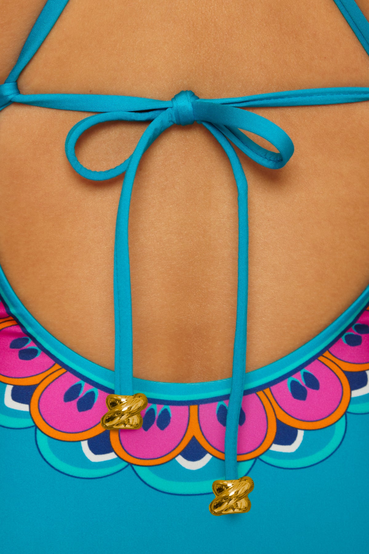 Close-up of the Mykonos Lace Up Maillot swimsuit back, showing blue straps tied in a bow with gold tips and a colorful scalloped neckline against tan skin.