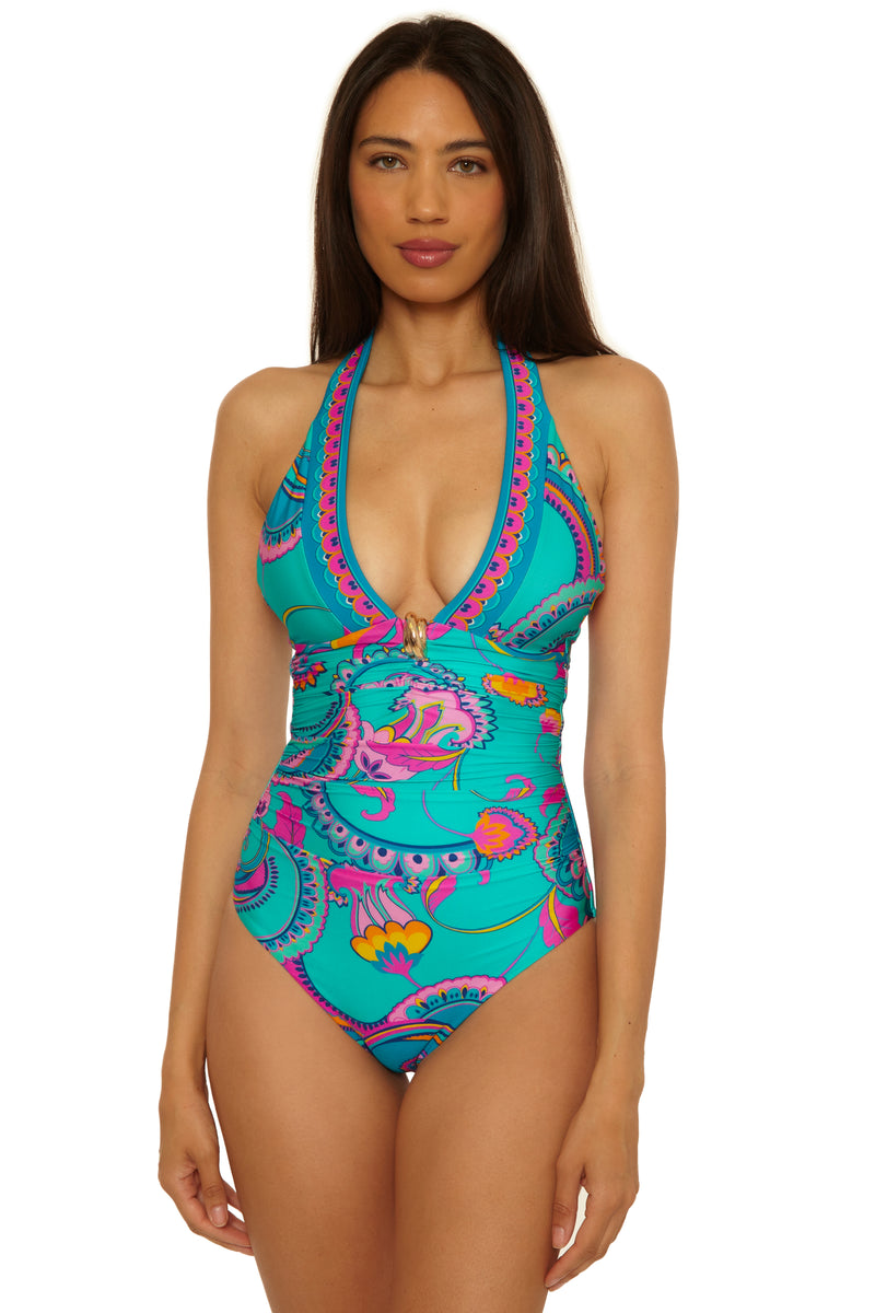 A woman models the Mykonos Plunge 1Pc, a halter-neck one-piece swimsuit with a vibrant turquoise, pink, and yellow paisley pattern and a deep V neckline, standing against a white background.
