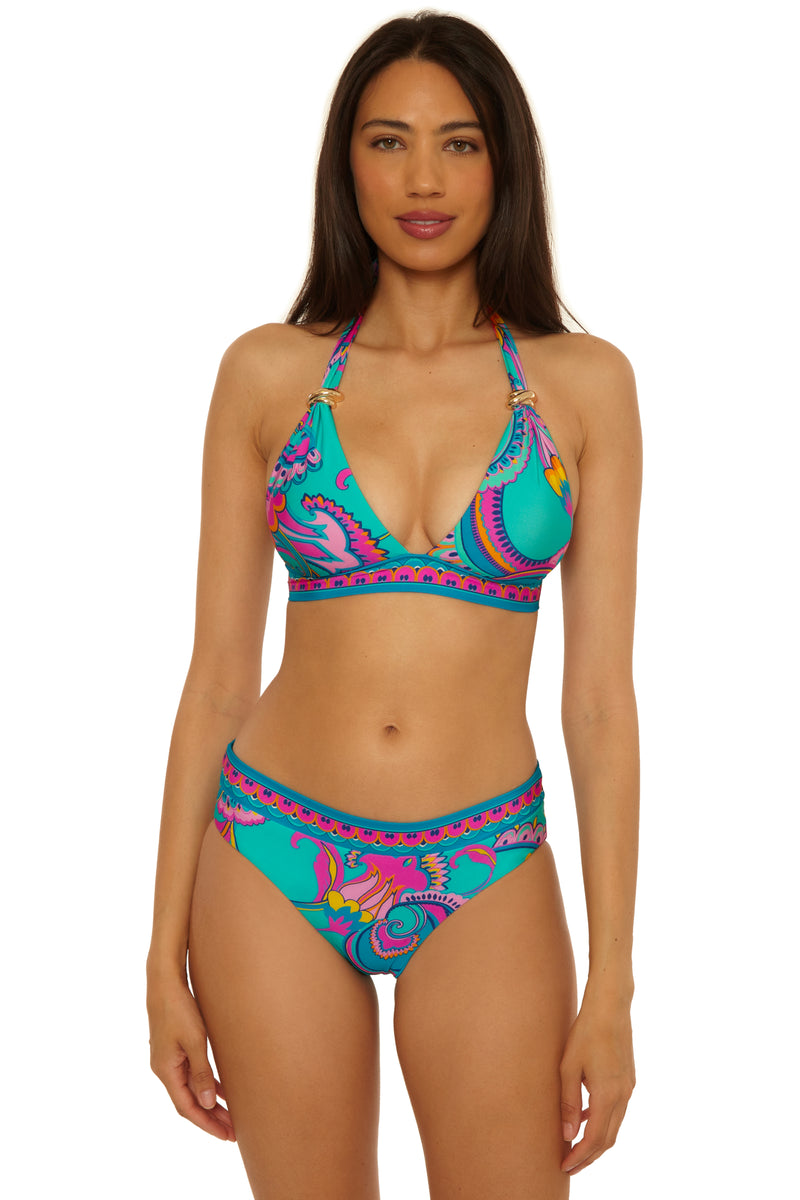 A woman with long dark hair wears the Mykonos Reversible Hipster bikini in a vibrant turquoise, pink, purple, and yellow pattern, standing facing forward with a neutral expression against a white background.