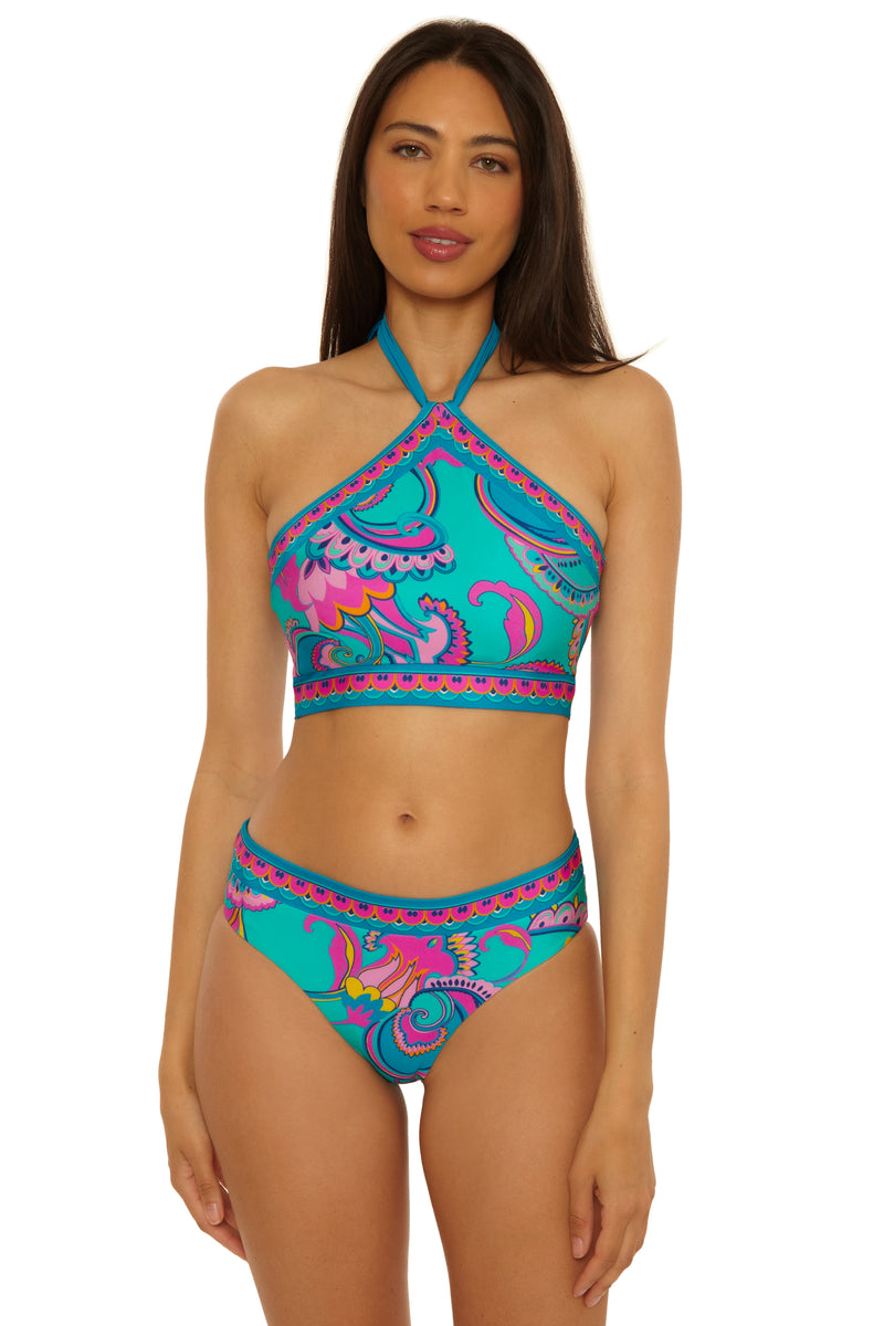 A woman models the Mykonos Reversible Hi Neck Top, a turquoise halter bikini featuring pink, purple, and blue paisley patterns, as she stands facing forward against a plain white background.