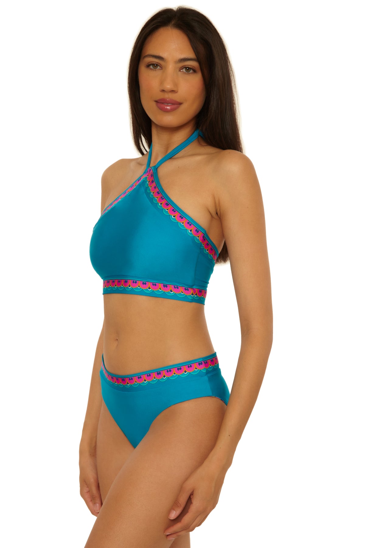 A woman models the Mykonos Reversible Hi Neck Top, a blue bikini with pink and patterned trim, posing against a white background.
