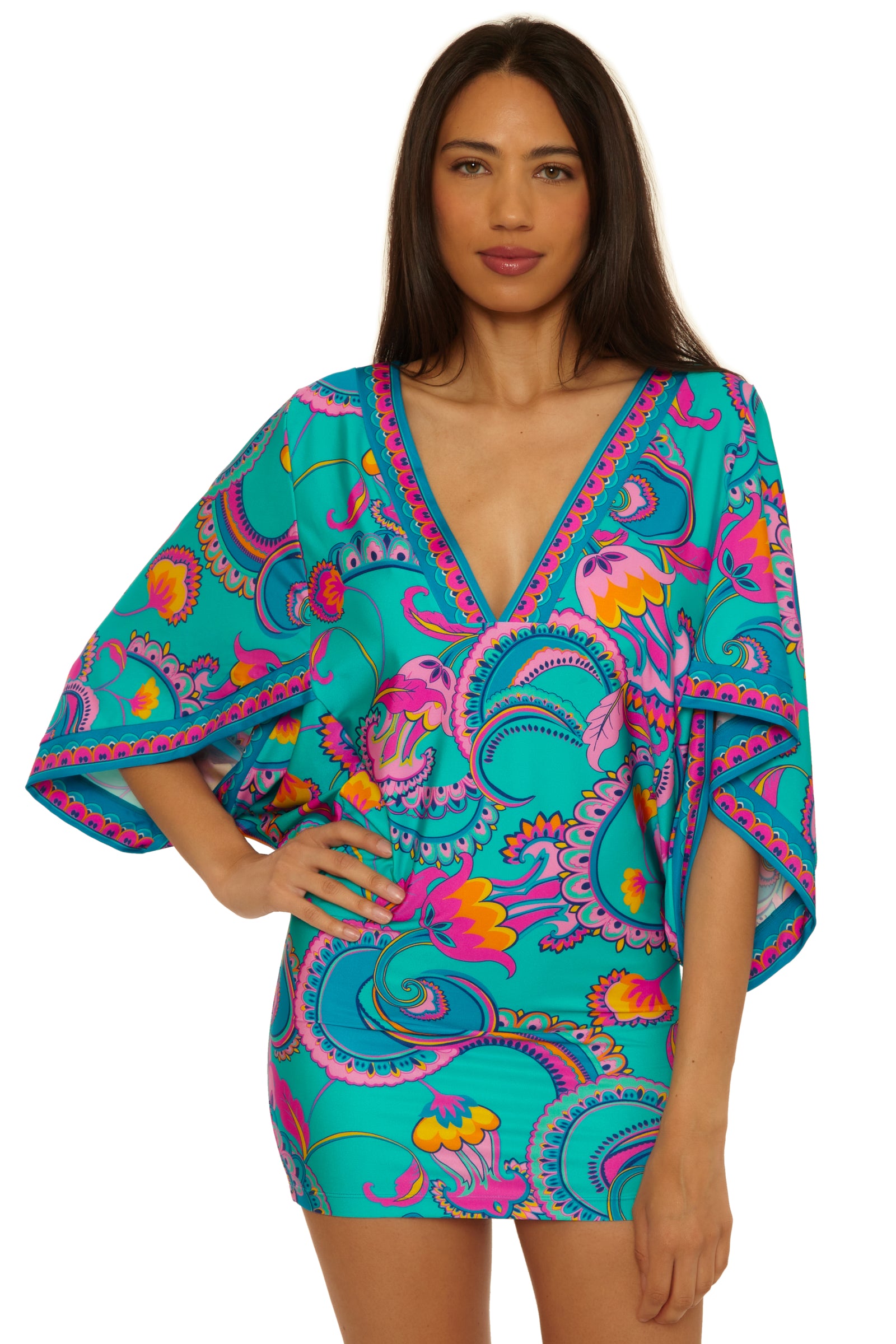 A woman wears the Mykonos Casablanca Tunic—a turquoise mini dress with a colorful paisley and floral print, wide sleeves, and a deep V-neckline. She has long brown hair and stands with one hand on her hip.