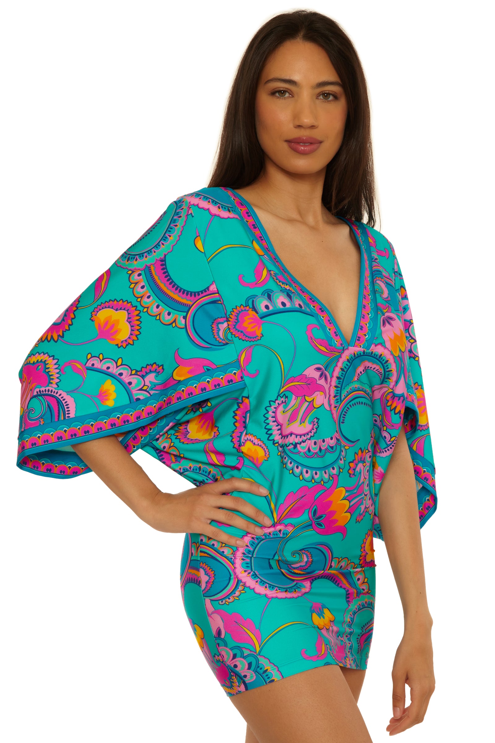 A woman wearing the Mykonos Casablanca Tunic, a colorful, patterned turquoise dress with pink, yellow, and purple swirls, stands with one hand on her hip and looks at the camera.