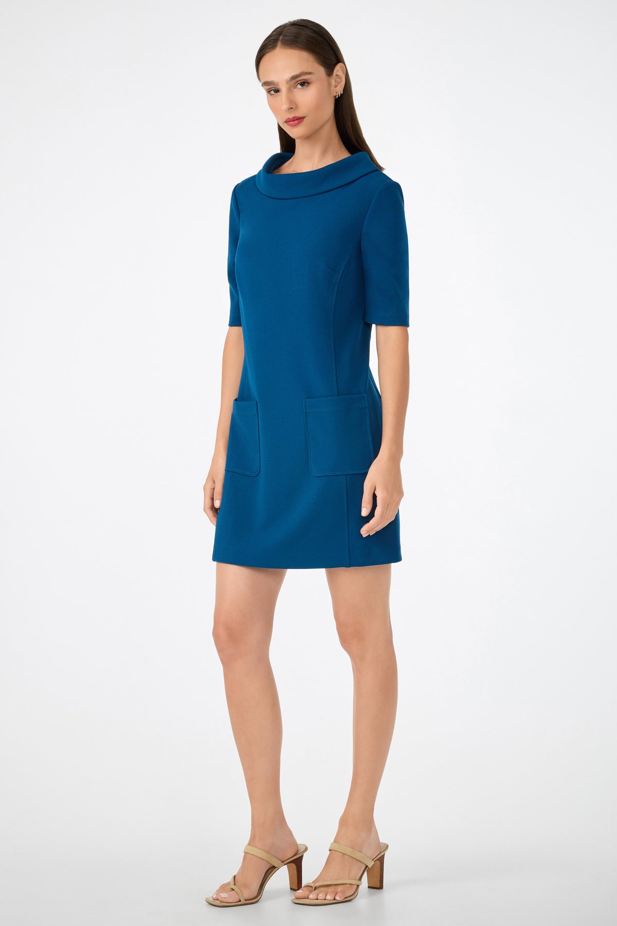 A woman with straight brown hair and a neutral expression stands against a plain background wearing the Maleko Dress—a teal, short-sleeved, knee-length dress with two front pockets—and beige heeled sandals.