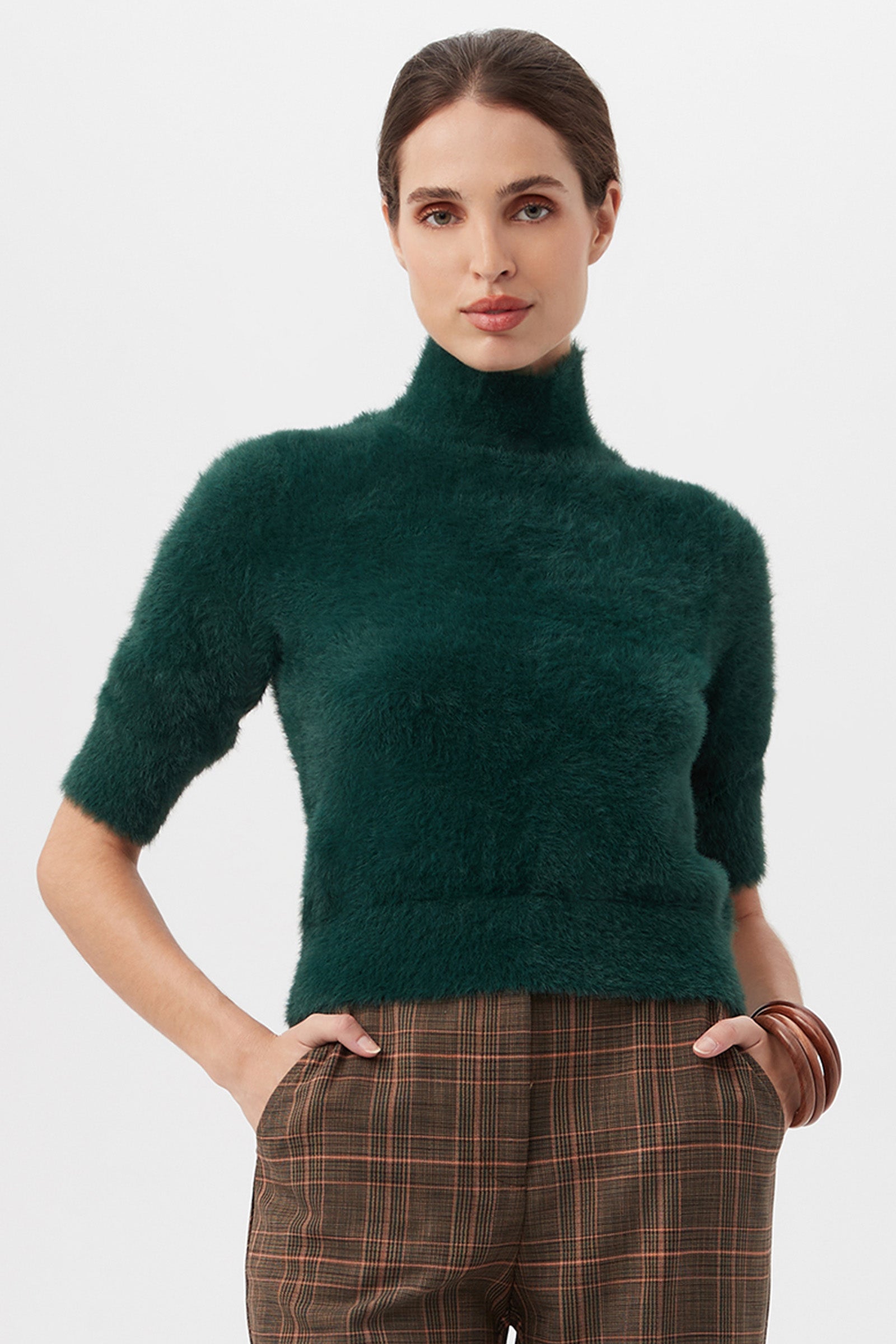 A woman wears the Mali Fuzzy Sweater in dark green with short sleeves, paired with brown plaid trousers, standing hands-in-pockets against a plain white background.