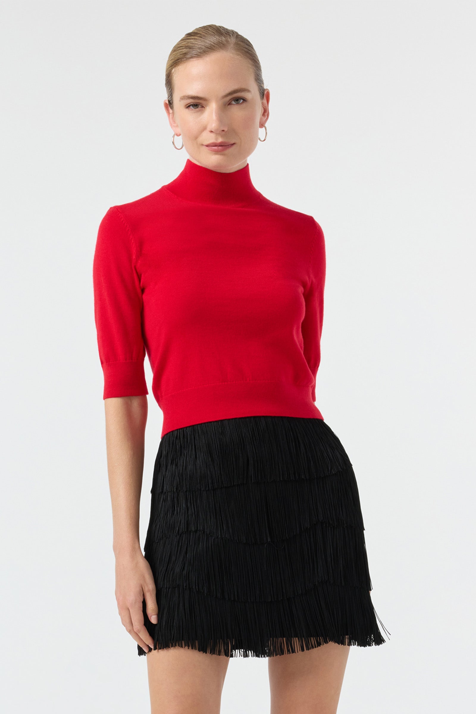 A woman with light hair pulled back stands against a plain background, confidently wearing the red Mali Mock Neck Pullover and a black, tiered fringe mini skirt.
