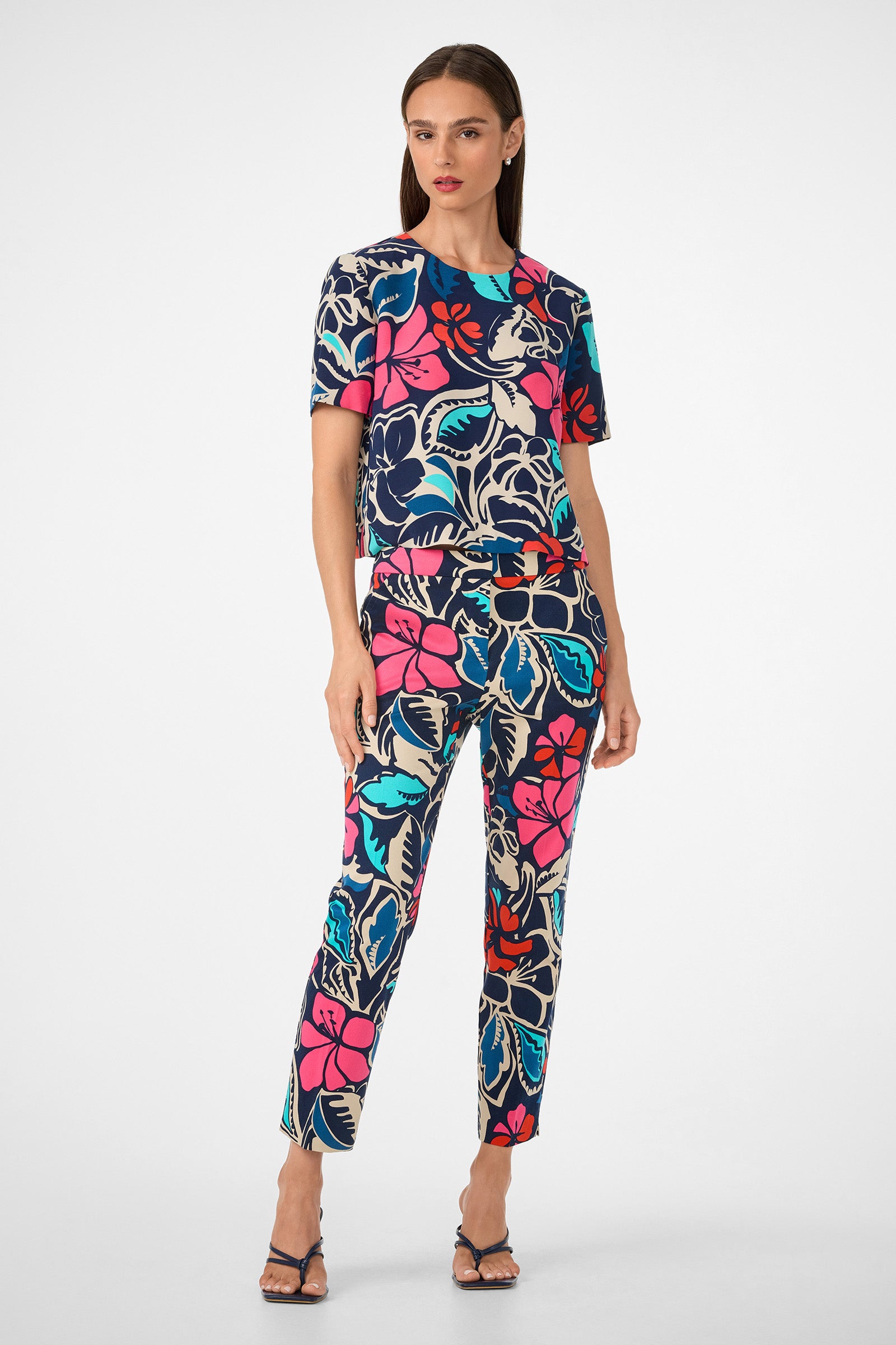 A woman with straight hair wears the Moss 2 Pant—a matching set featuring a bold floral print in pink, red, blue, black, and cream—paired with a short-sleeve top and black heeled sandals against a plain background.