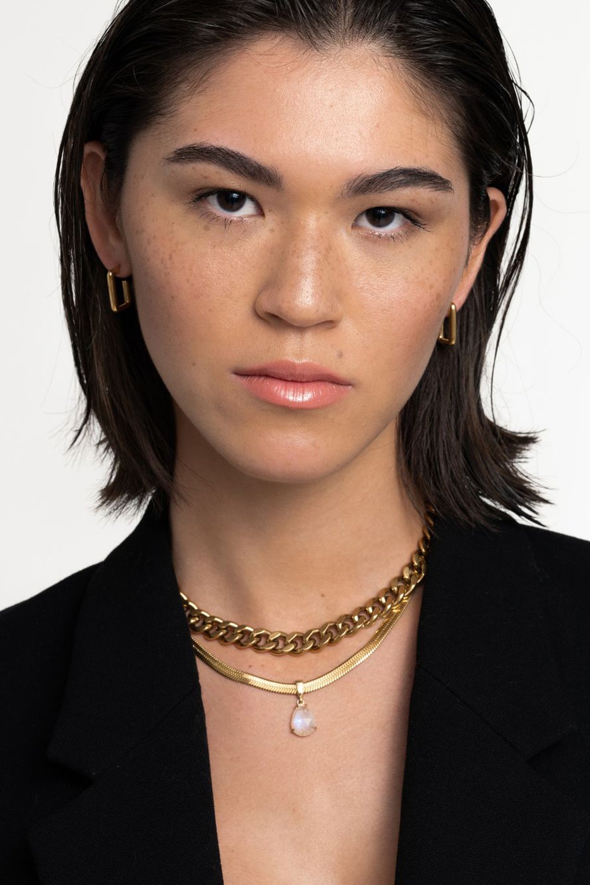 A woman with short brown hair wears gold hoop earrings and layered Alize Necklaces with a black blazer. She has a neutral expression, natural makeup, and poses against a plain white background.