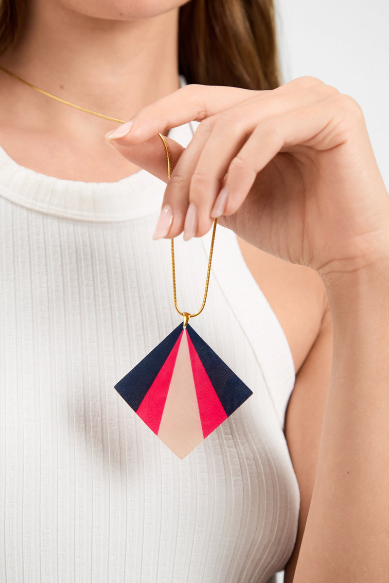 Wearing a white sleeveless top, a person holds the Kite Necklace, featuring geometric navy blue, red, and beige color blocks on a pendant suspended from a thin gold chain.