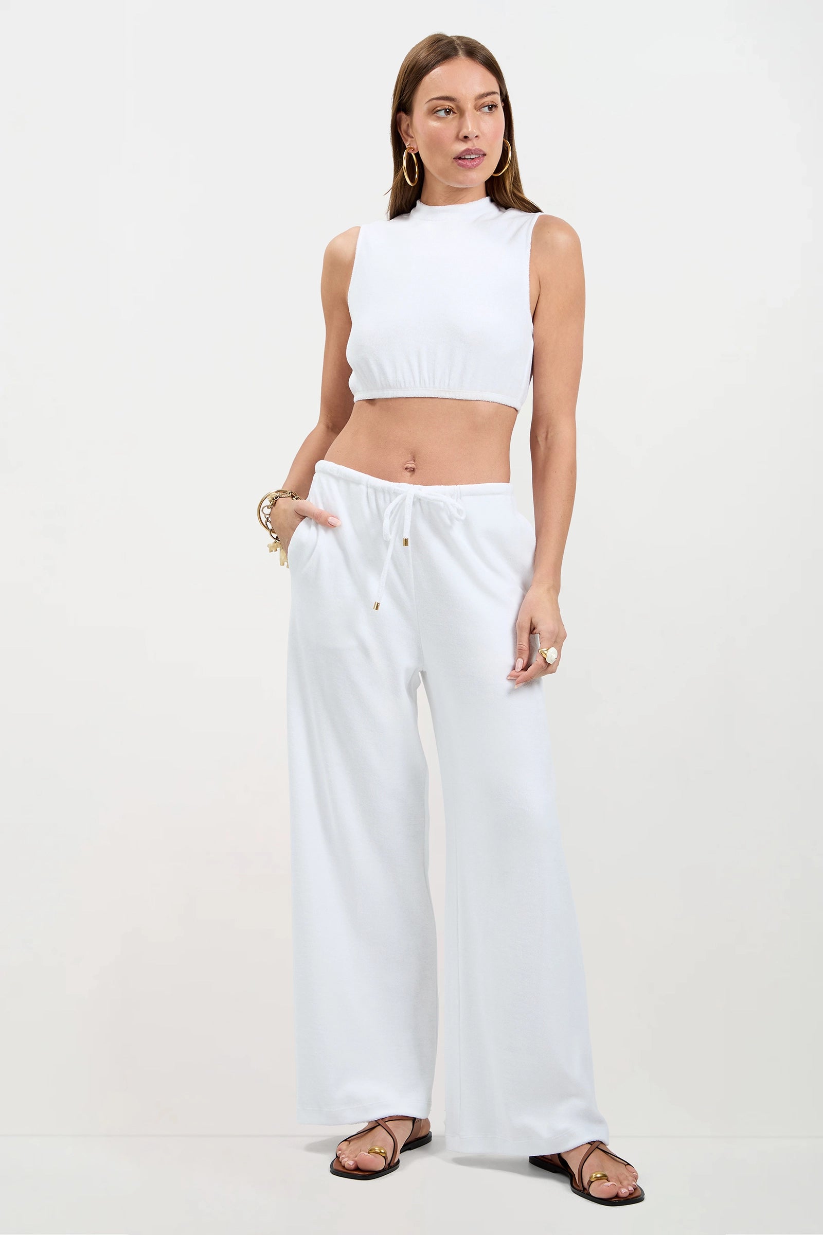 A woman stands against a plain background wearing the Nelda Sleeveless Crop Top - Poolside Terry in white and matching wide-leg pants. She accessorizes with hoop earrings, bracelets, rings, sandals, and has one hand in her pocket.