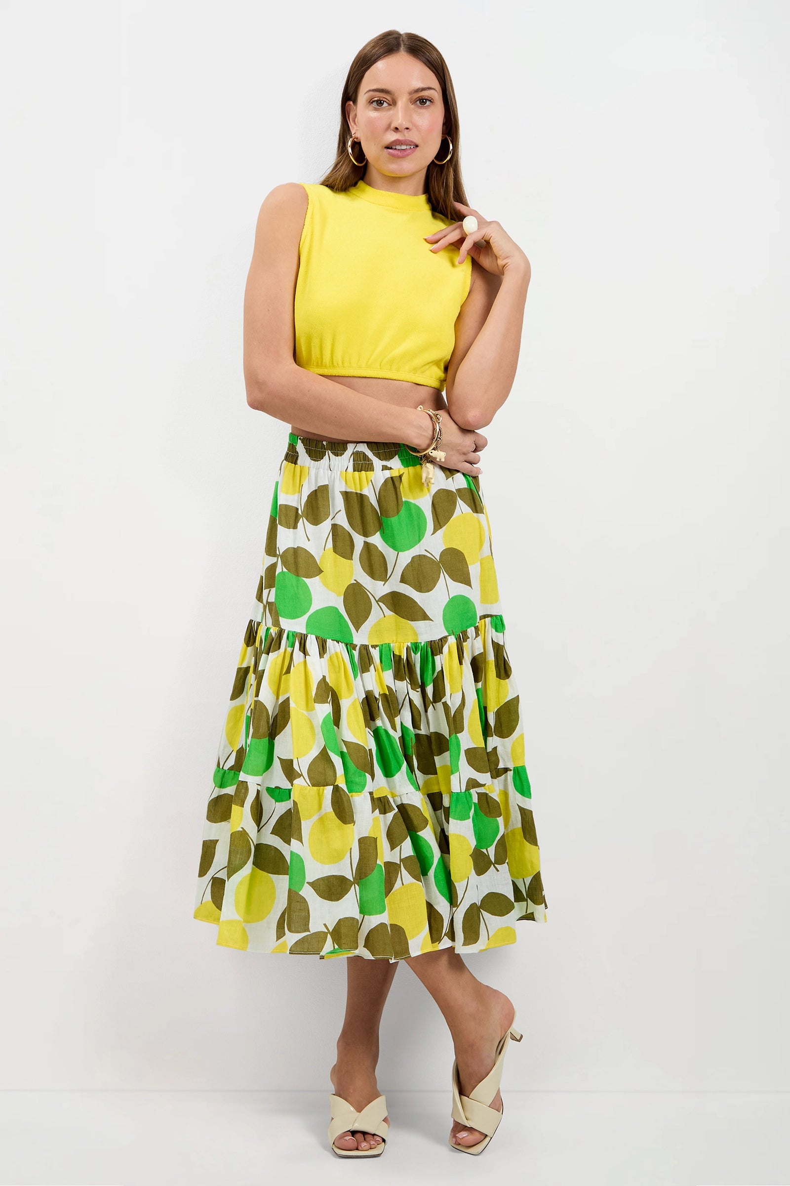 A woman stands against a white wall wearing the Nelda Sleeveless Crop Top - Poolside Terry and a tiered, ankle-length skirt with a green and yellow leaf pattern. She has long hair, gold hoop earrings, and beige heeled sandals.