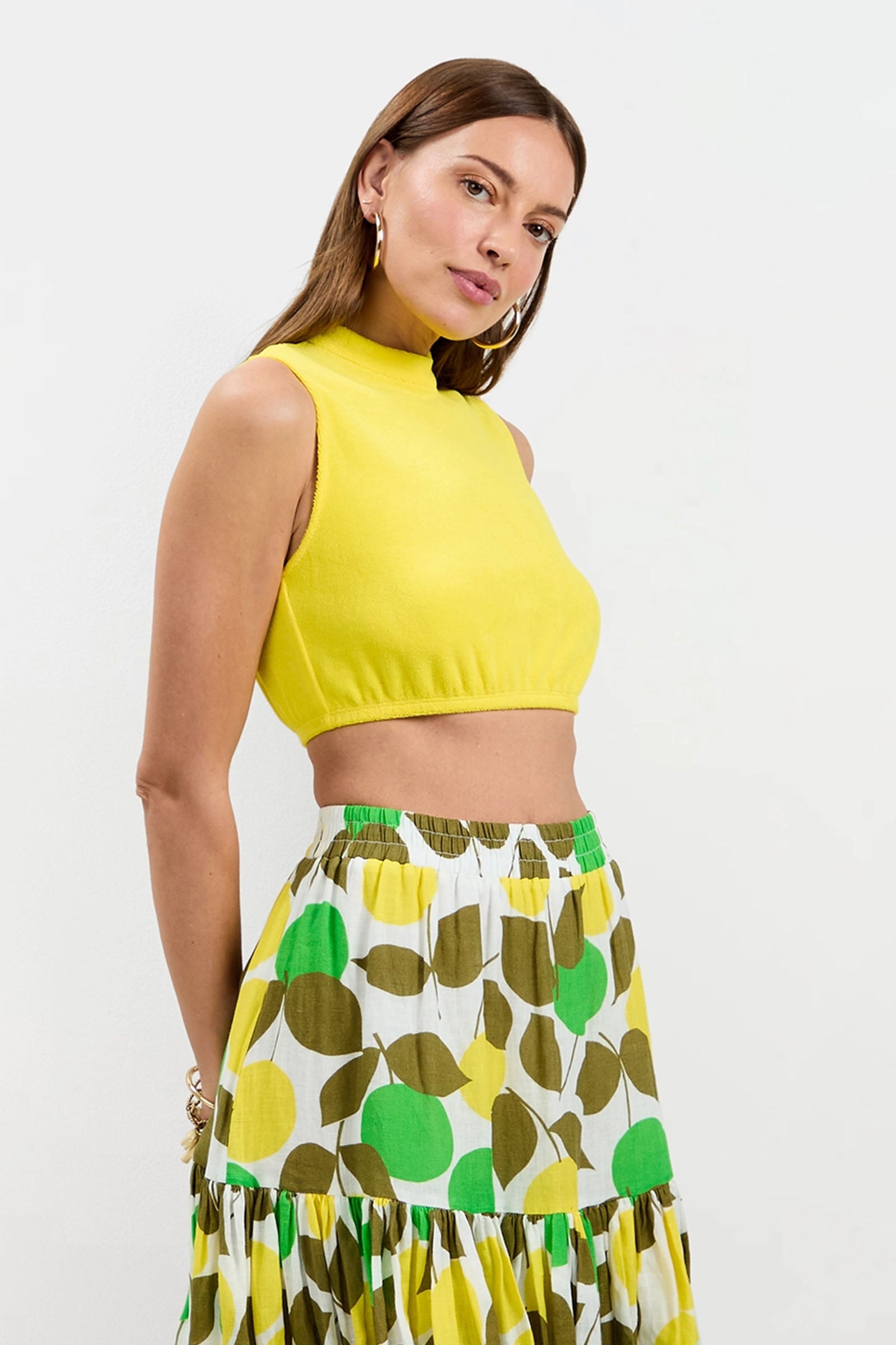 A woman wears the Nelda Sleeveless Crop Top - Poolside Terry and a high-waisted white skirt with green and olive leaf patterns, standing against a plain white background.