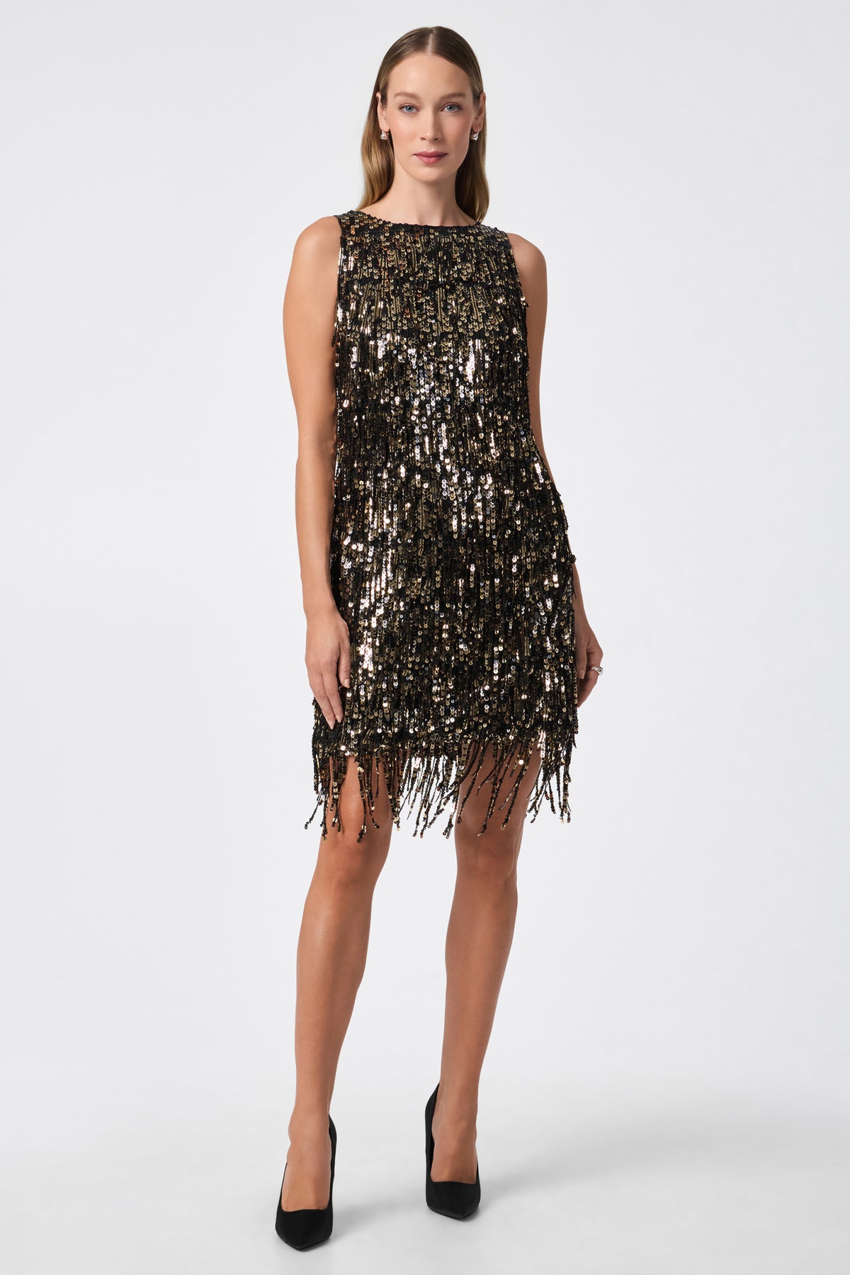 A woman stands against a white background wearing the Nini Dress in BLACK/GOLD, a sleeveless, knee-length dress with shimmering sequins and fringe, paired with black high heels.