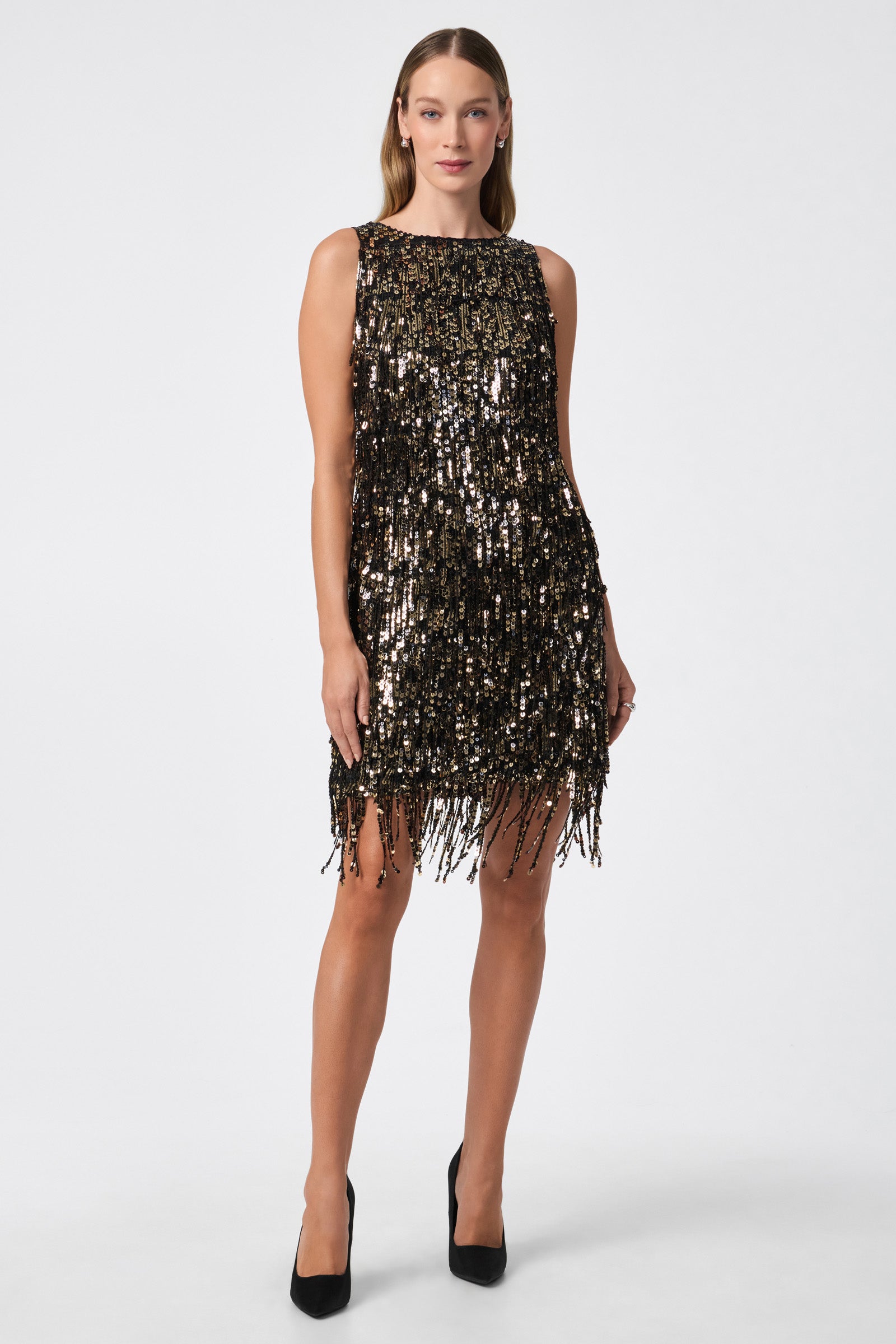 A woman stands against a white background wearing the Nini Dress in BLACK/GOLD, a sleeveless, knee-length dress with shimmering sequins and fringe, paired with black high heels.