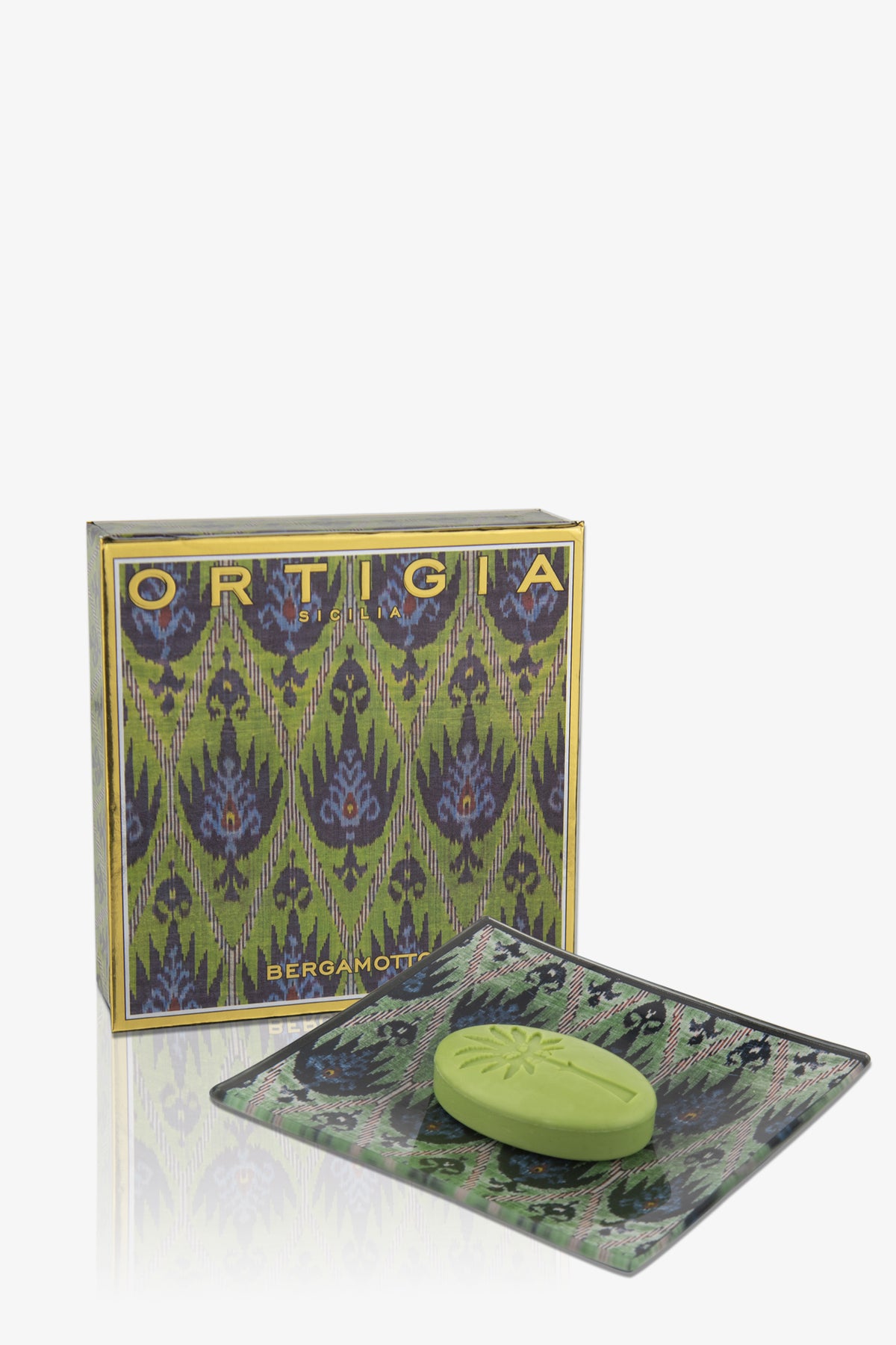 A green Ortigia Bergamot Soap Plate rests on a decorative glass dish in front of a patterned box with blue and gold accents.