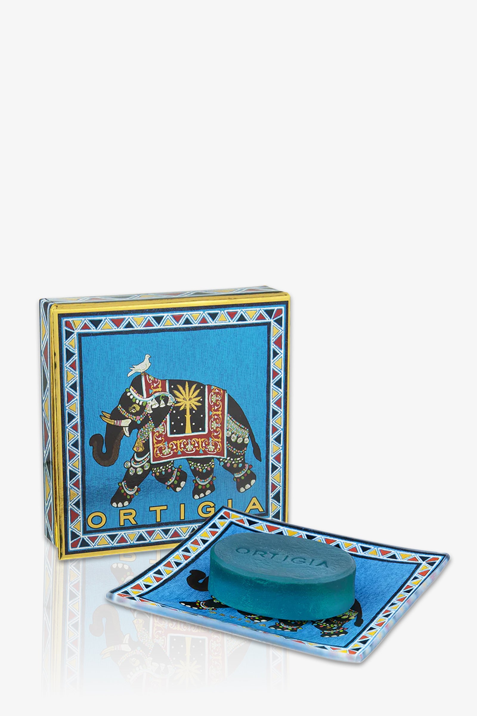 The Ortigia Sandalo Soap Plate Set in blue features a soap bar on a matching decorative tray, with a blue box adorned by a colorful, ornate elephant illustration in the background.