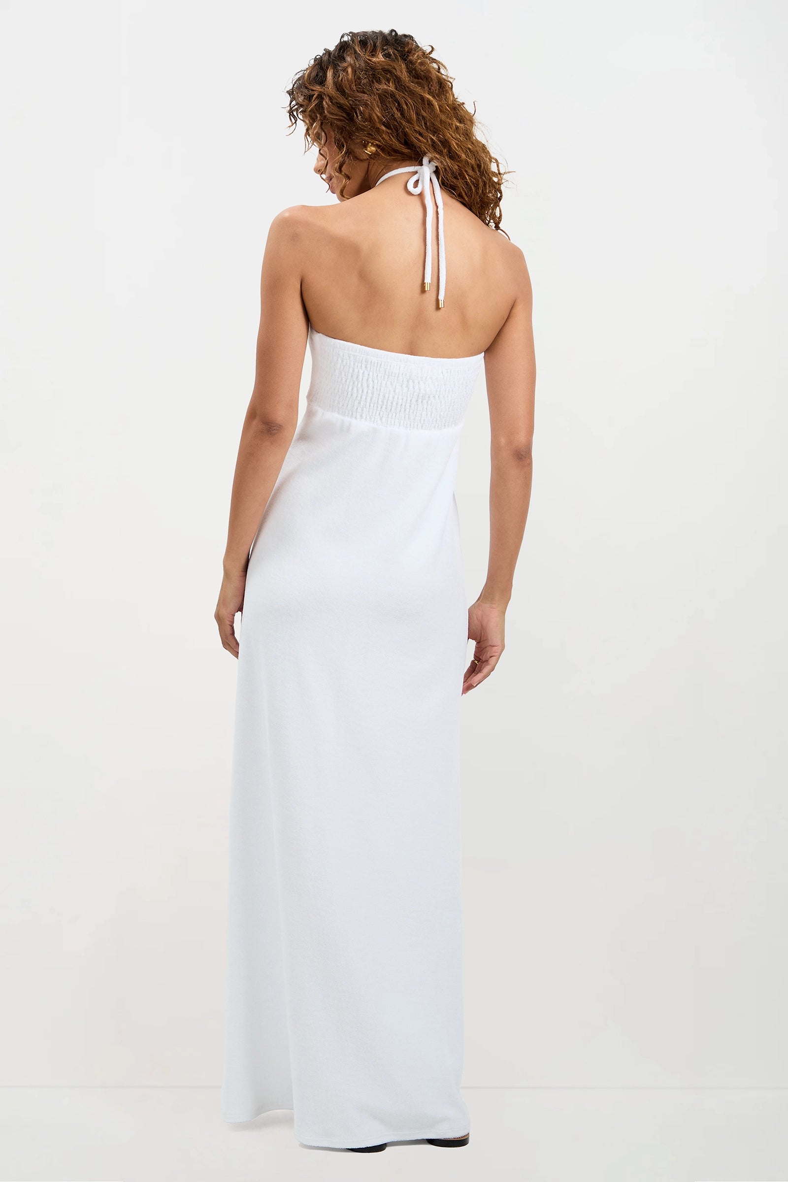 A woman with curly hair stands facing away, wearing the Palm Sleeveless Maxi Dress - Poolside Terry, a long white strapless dress with a halter neck tie and elastic back, against a plain light-colored background.