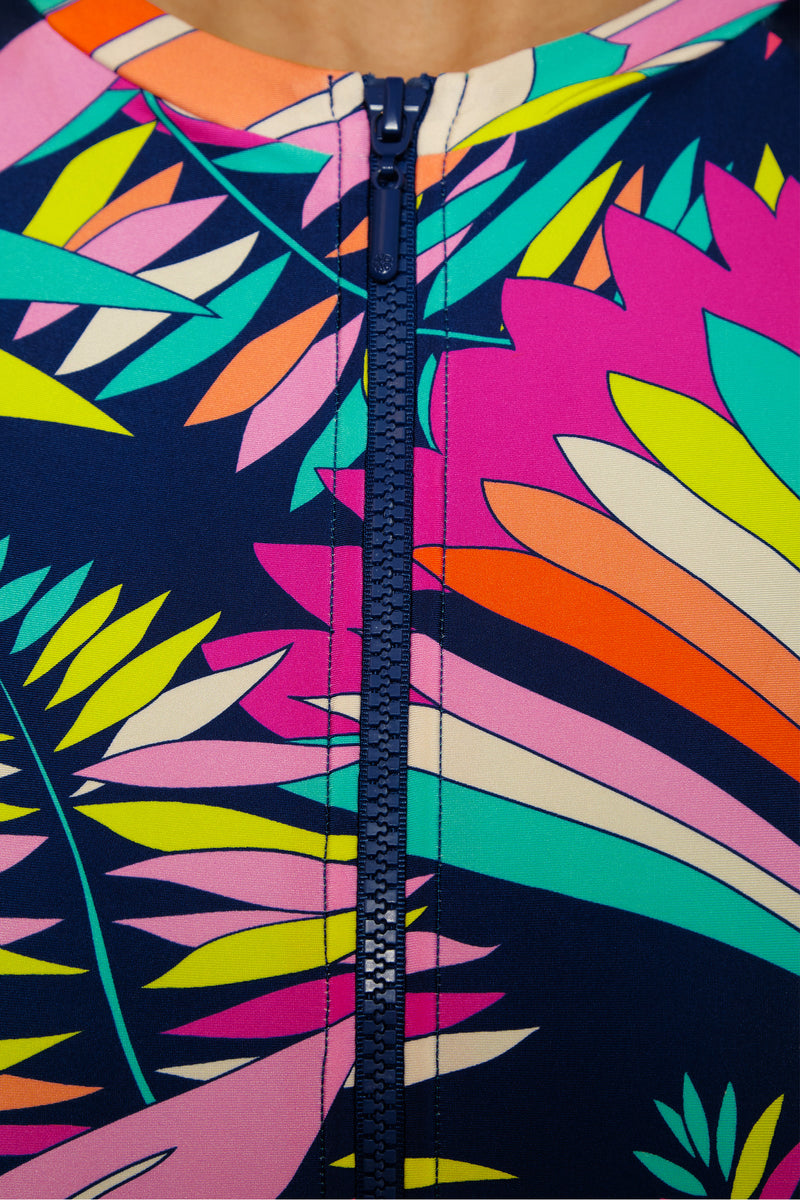 Close-up of the Paralia Zip Up Paddlesuit featuring a blue zipper and a vibrant tropical leaf pattern in pink, yellow, green, orange, and blue on a dark background.