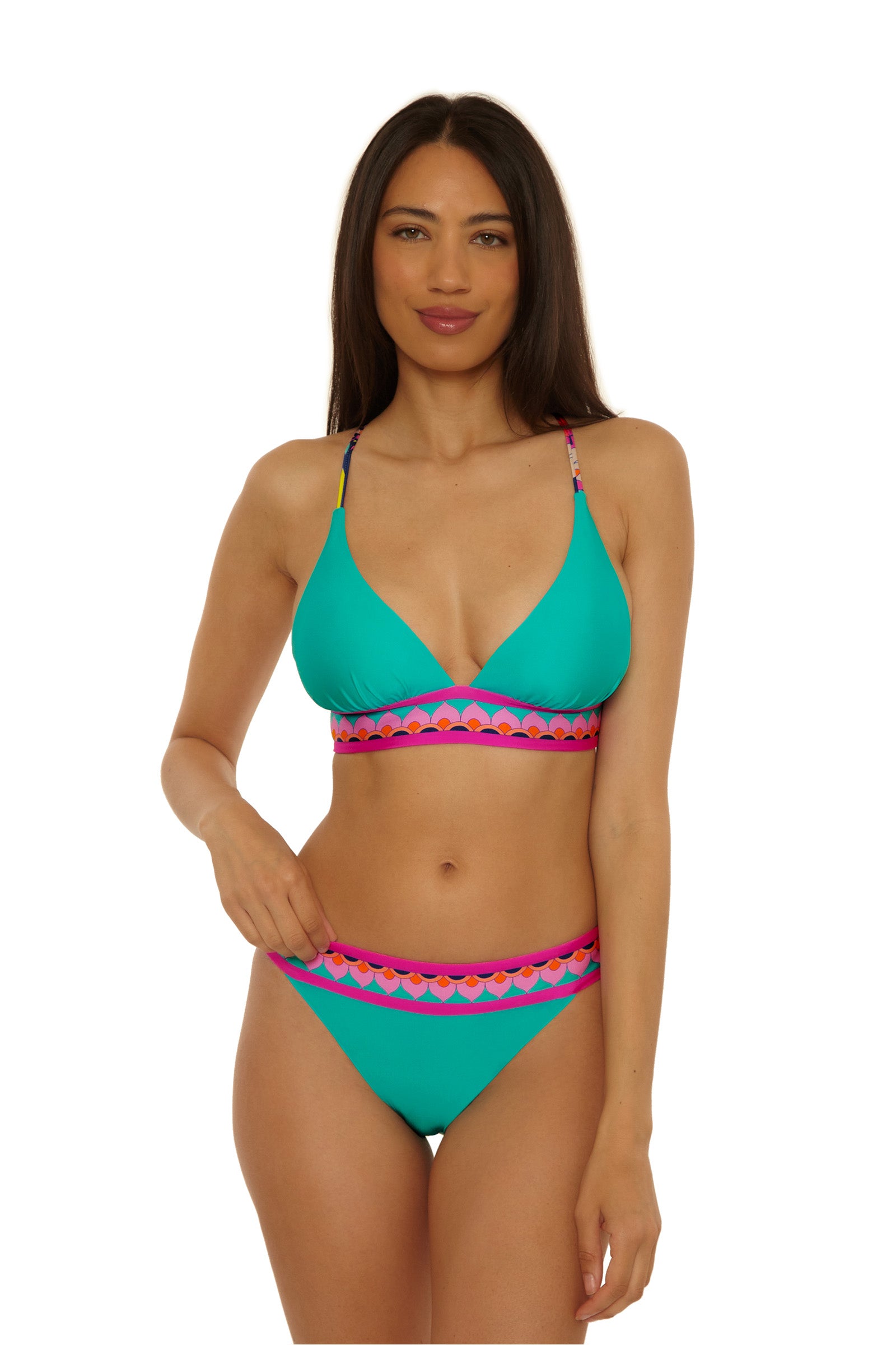 A woman with long brown hair and a slight smile wears the Paralia Reversible Halter Top in teal with pink and purple scalloped trim, standing against a plain white background with one hand on her hip.