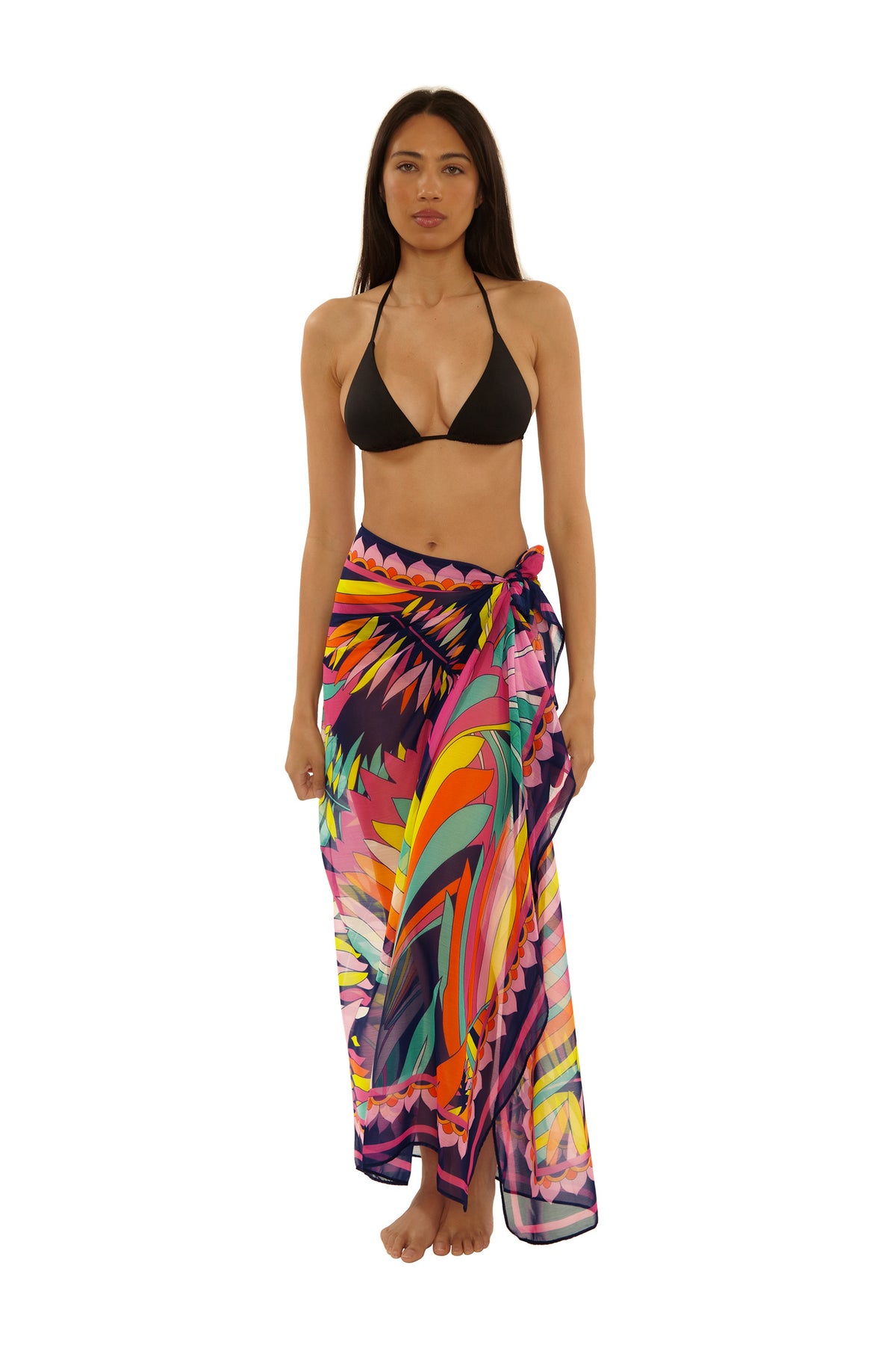 A woman stands barefoot in a black bikini top and the Paralia Pareo, a colorful tropical sarong tied at the waist, against a plain white background.
