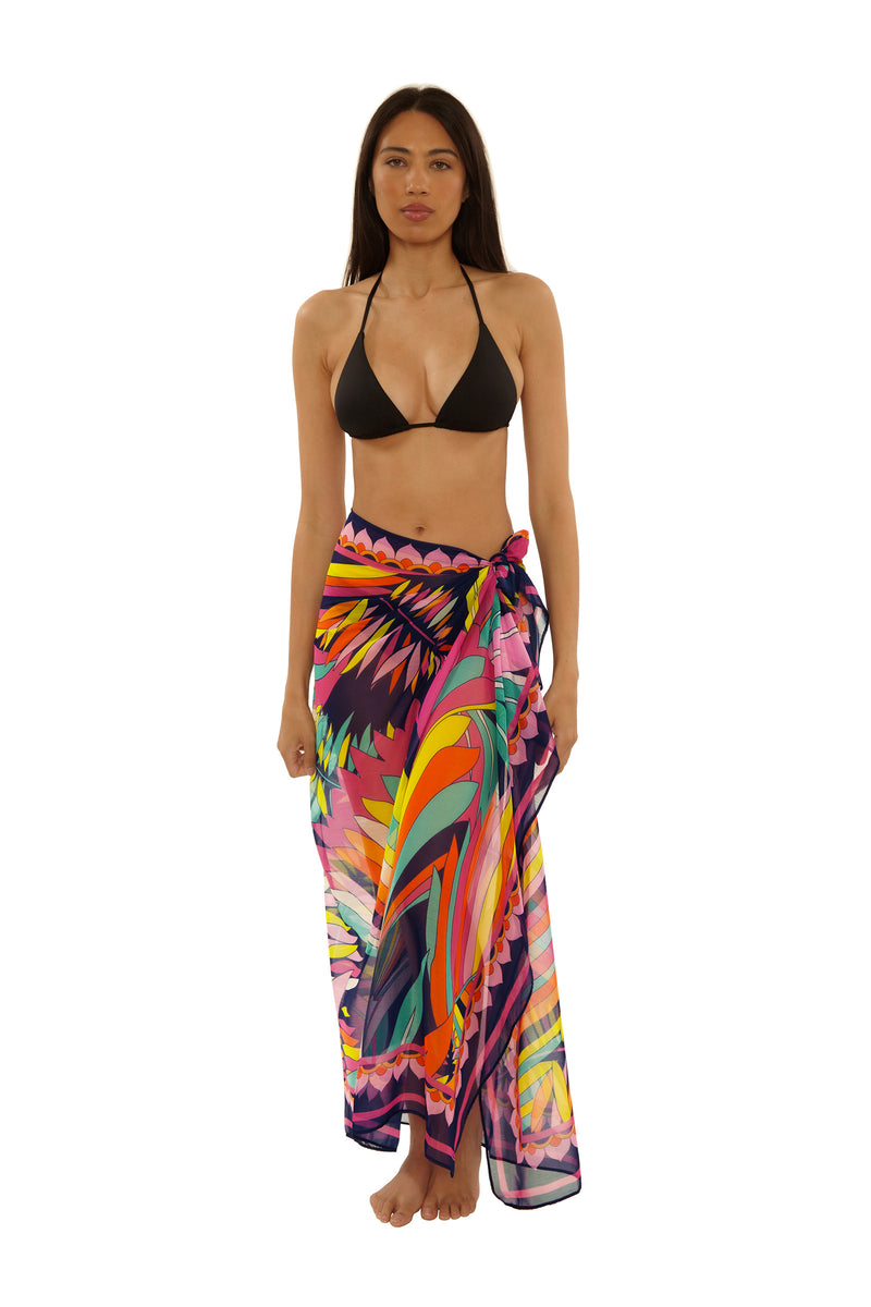 A woman stands barefoot in a black bikini top and the Paralia Pareo, a colorful tropical sarong tied at the waist, against a plain white background.