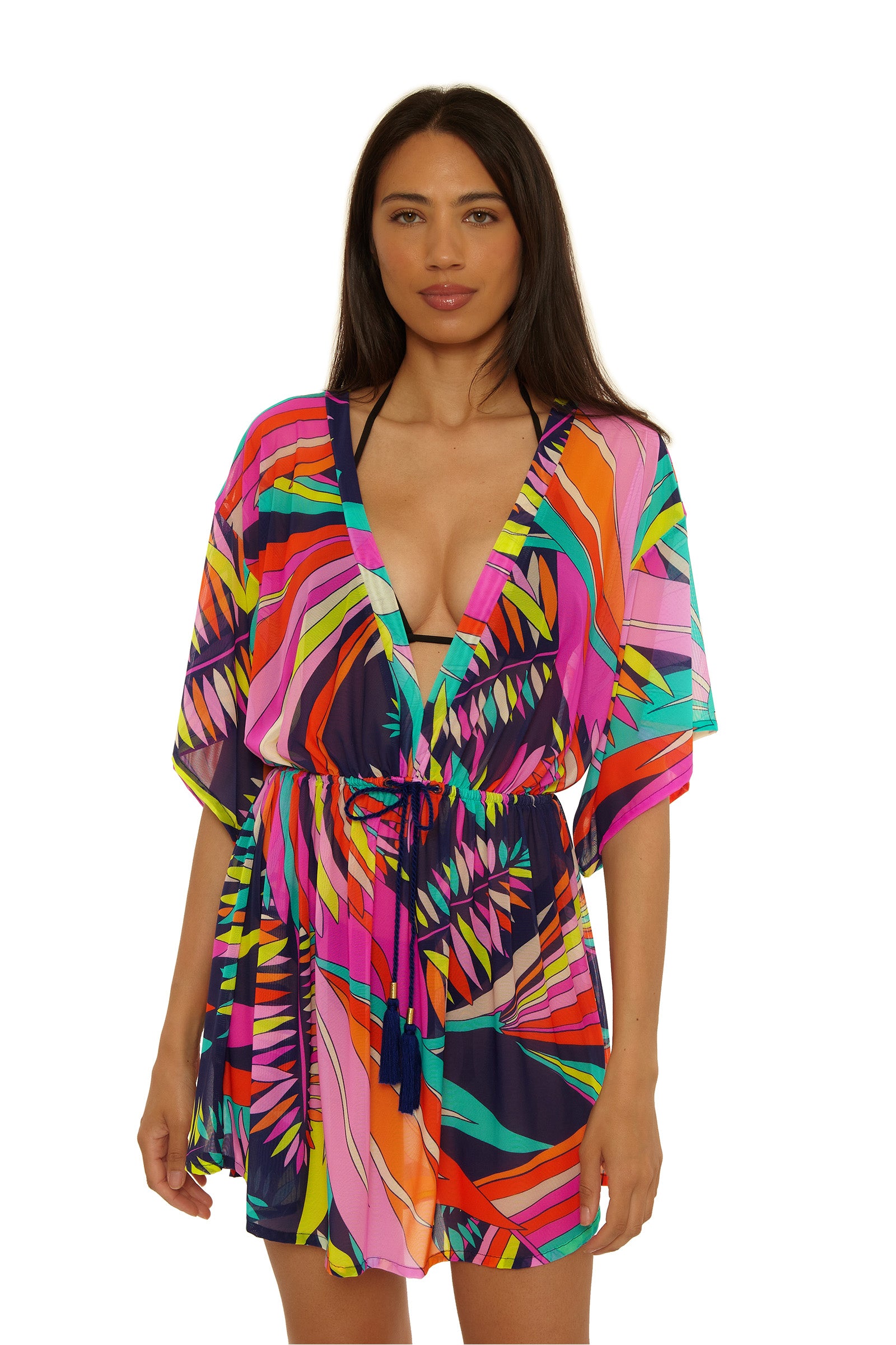 A woman with long dark hair wears the Paralia Mesh Tunic Dress—a colorful, abstract-patterned dress with a deep V-neck, short sleeves, and drawstring waist—standing against a plain white background.