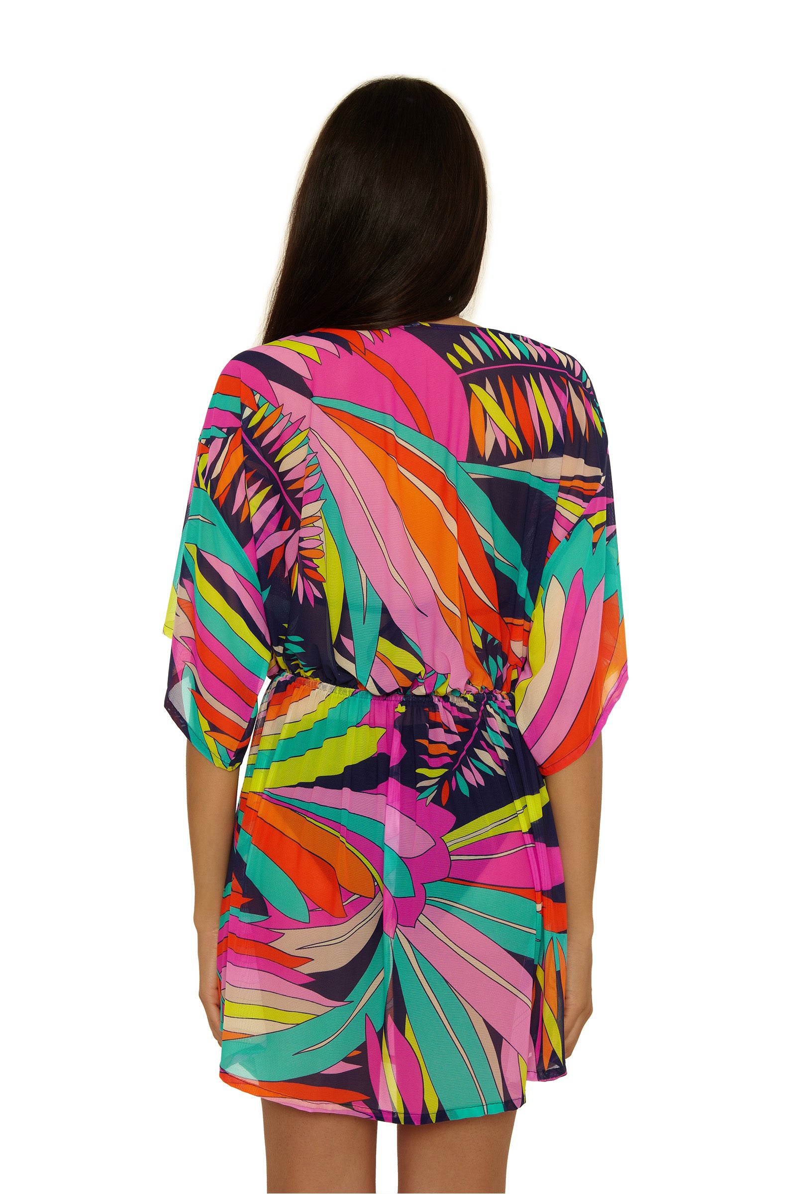 A woman with long dark hair stands with her back to the camera, wearing the Paralia Mesh Tunic Dress—a short, loose dress featuring brightly colored abstract leaf patterns in pink, orange, yellow, green, and purple.