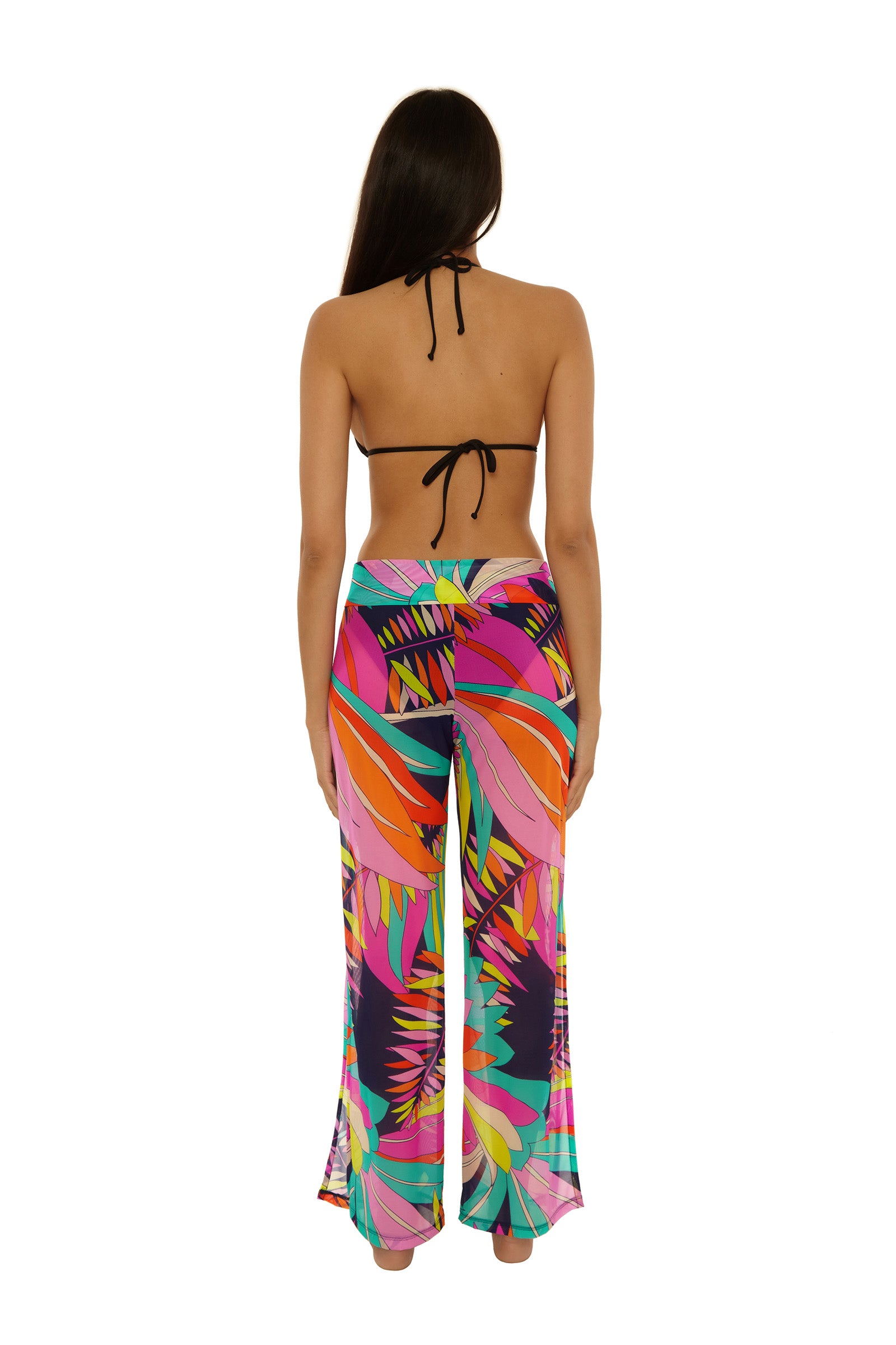 A woman with long brown hair, seen from the back, models the Paralia Mesh Swim Pant—a pair of brightly colored, tropical-patterned wide-leg pants—paired with a black halter bikini top against a plain white background.