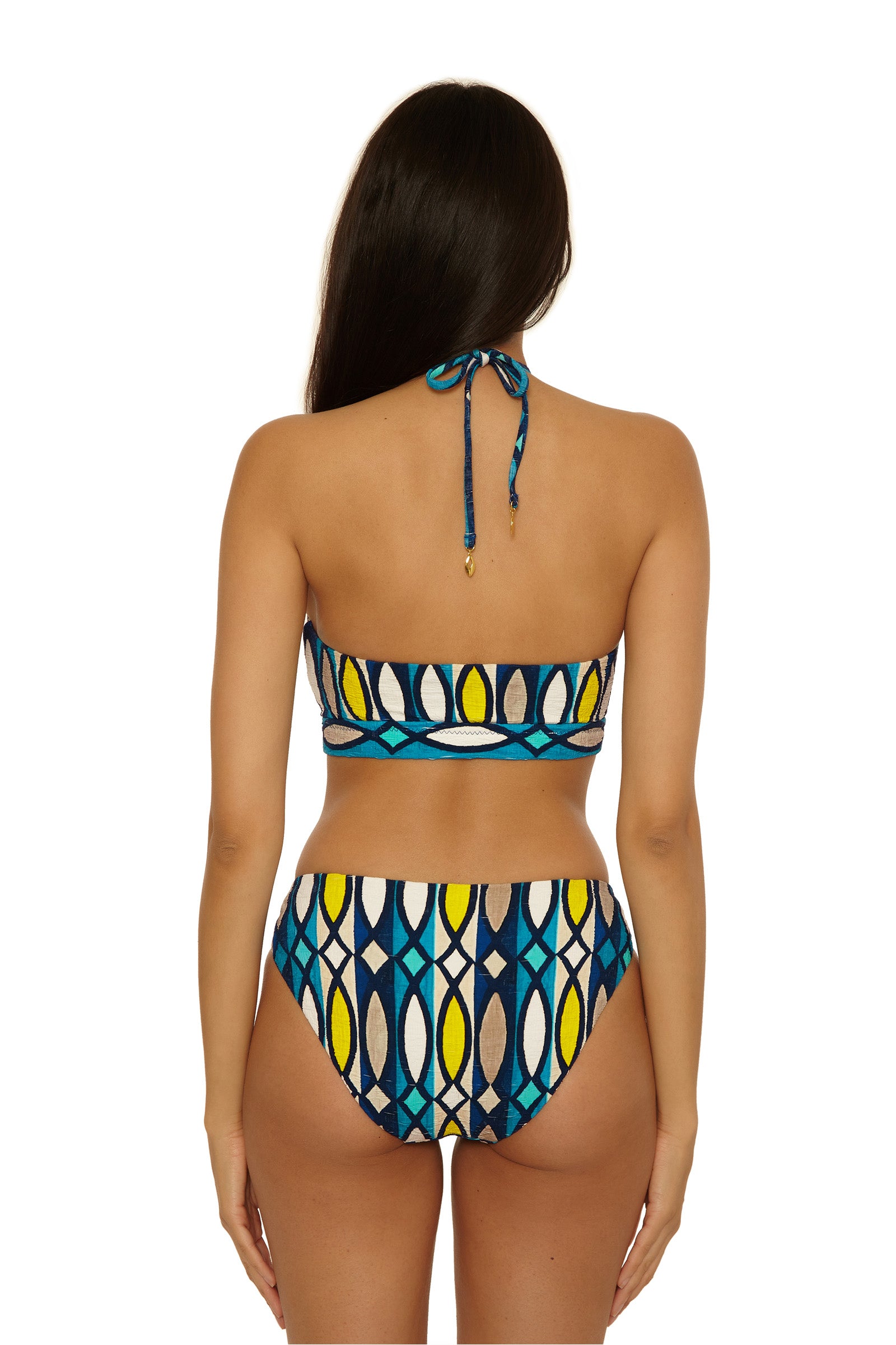 A woman with long dark hair is shown from the back, wearing the Paros Banded Bandeau Top featuring a blue, yellow, white, and black geometric pattern. The bikini top ties at the neck and is paired with high-waisted bottoms.