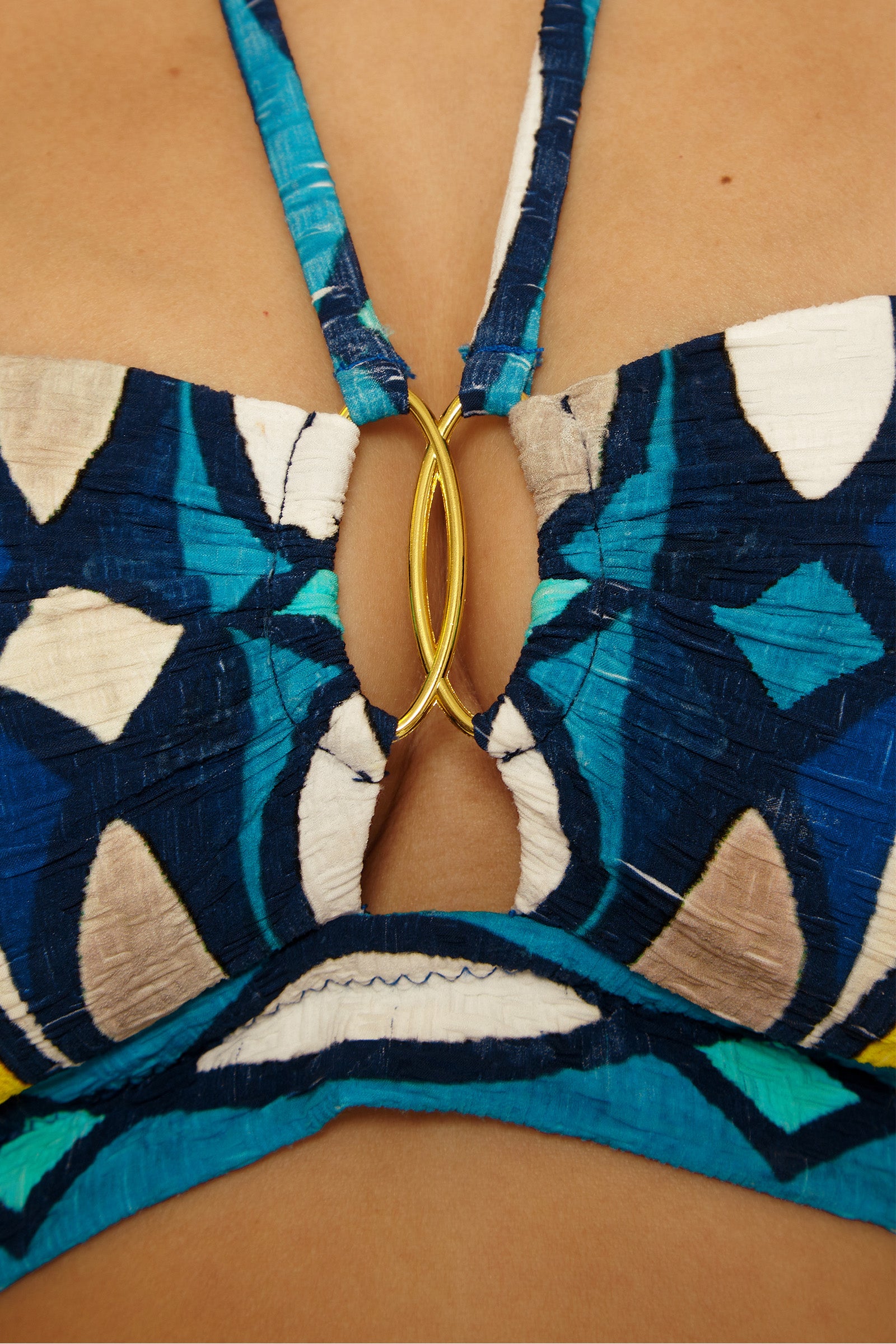 Close-up of a person wearing the Paros Banded Bandeau Top, featuring a blue, white, and teal pattern with a gold oval ring detail at the center of the chest.