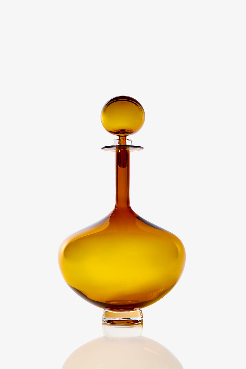 The Petite Genie Bottle Decanter features a round, amber glass design with a wide base, narrow neck, and matching spherical stopper, shown on a white background.