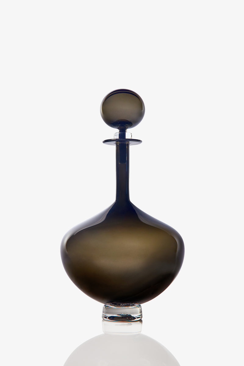 The Petite Genie Bottle Decanter features dark, smoky glass with a wide rounded base, a long narrow neck, and a spherical stopper, displayed against a white background.