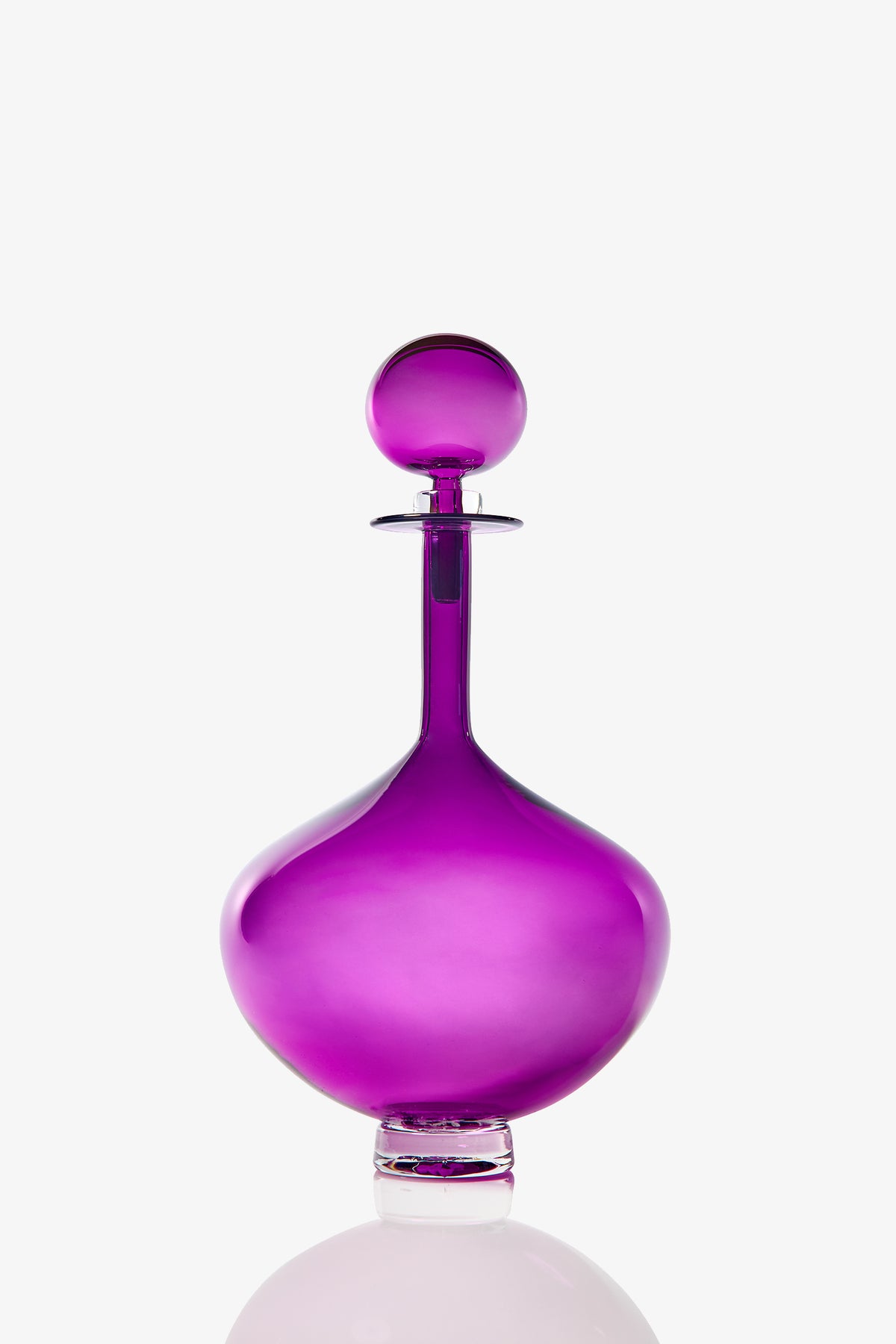 The Petite Genie Bottle Decanter is a round, elegant glass decanter with a wide base, narrow neck, and matching spherical stopper. Transparent and vibrant purple, it stands out against a plain white background.
