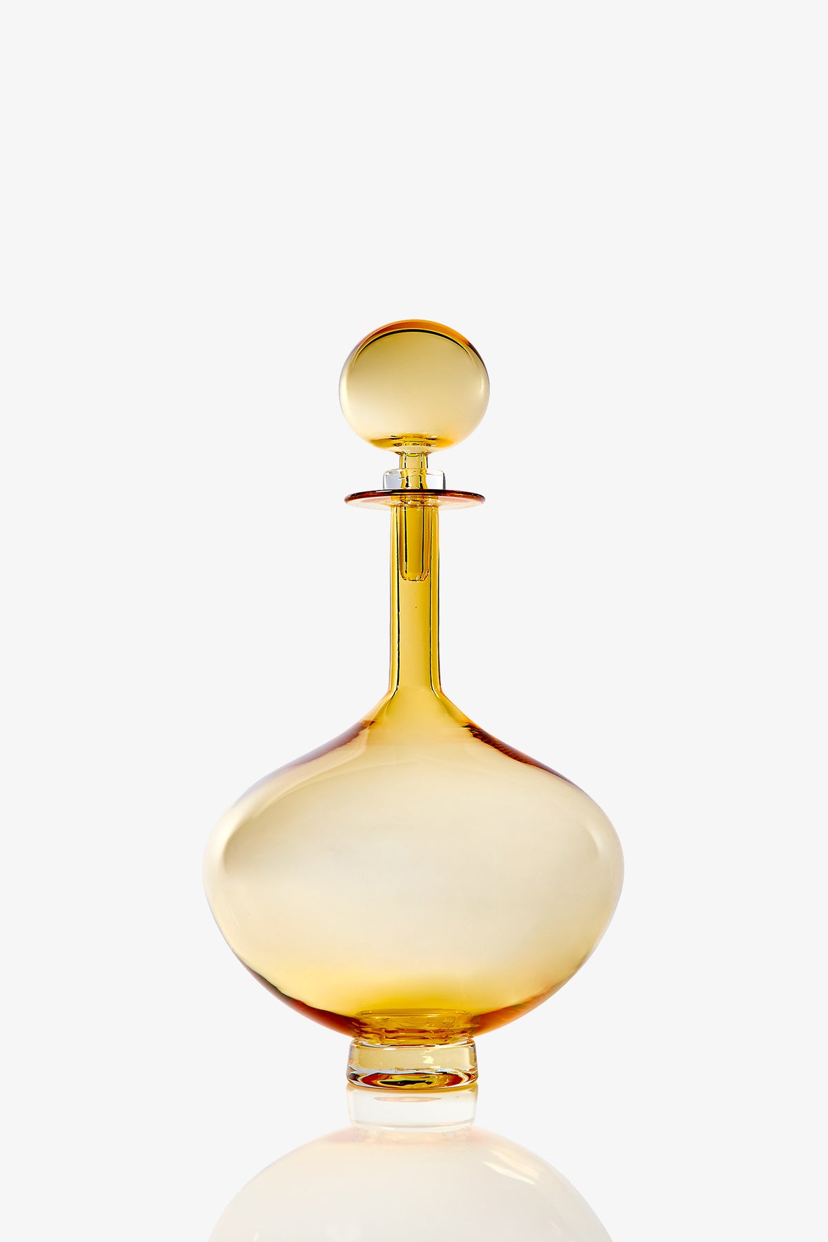 The Petite Genie Bottle Decanter features translucent amber glass with a round stopper, wide body, and short neck. It stands on a clear flat base and is set against a plain white background.