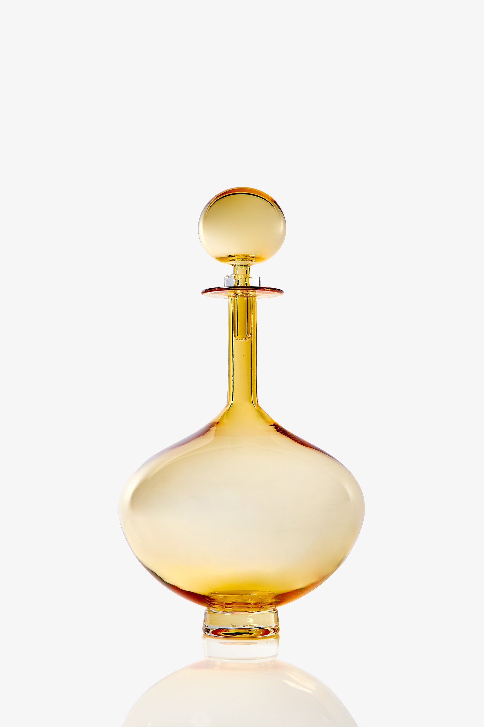 The Petite Genie Bottle Decanter features translucent amber glass with a round stopper, wide body, and short neck. It stands on a clear flat base and is set against a plain white background.