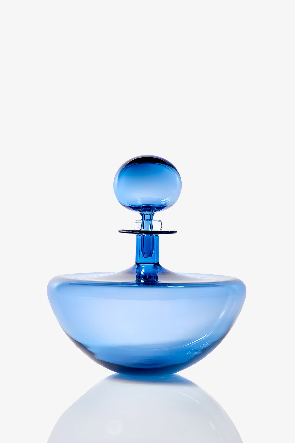The Small Angelic Bottle, a translucent blue glass perfume bottle with a round stopper, sits on a reflective surface against a white background.