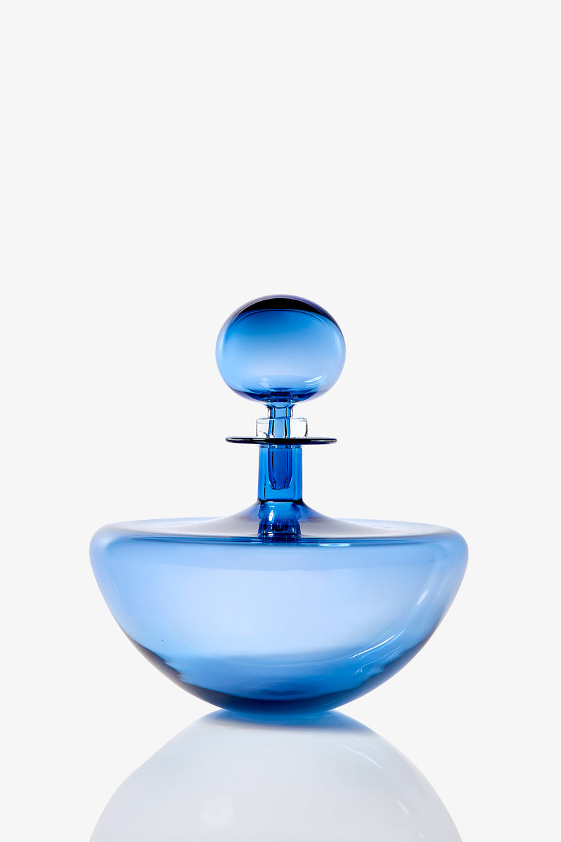 The Small Angelic Bottle, a translucent blue glass perfume bottle with a round stopper, sits on a reflective surface against a white background.