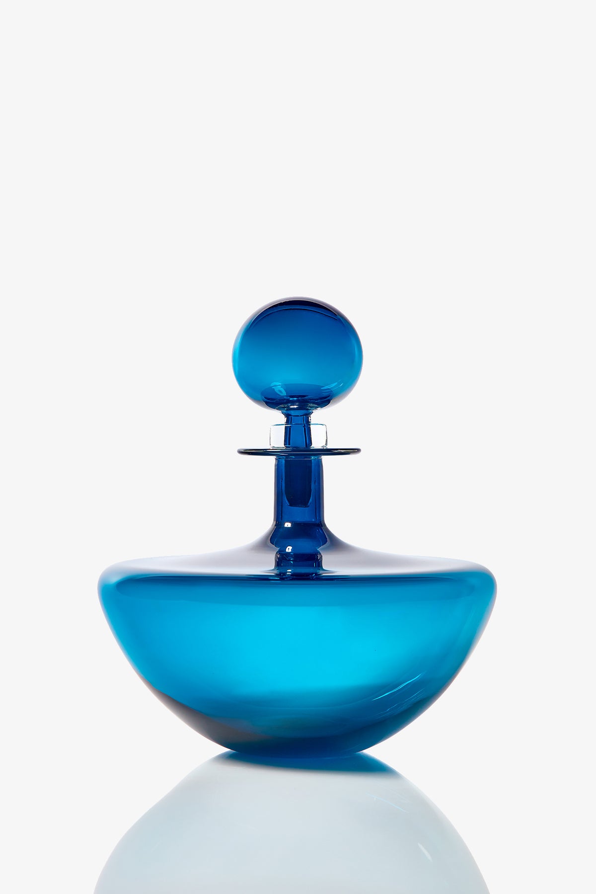 The Small Angelic Bottle is a sleek, modern blue glass perfume bottle with a round stopper, displayed on a reflective white surface against a simple white background.
