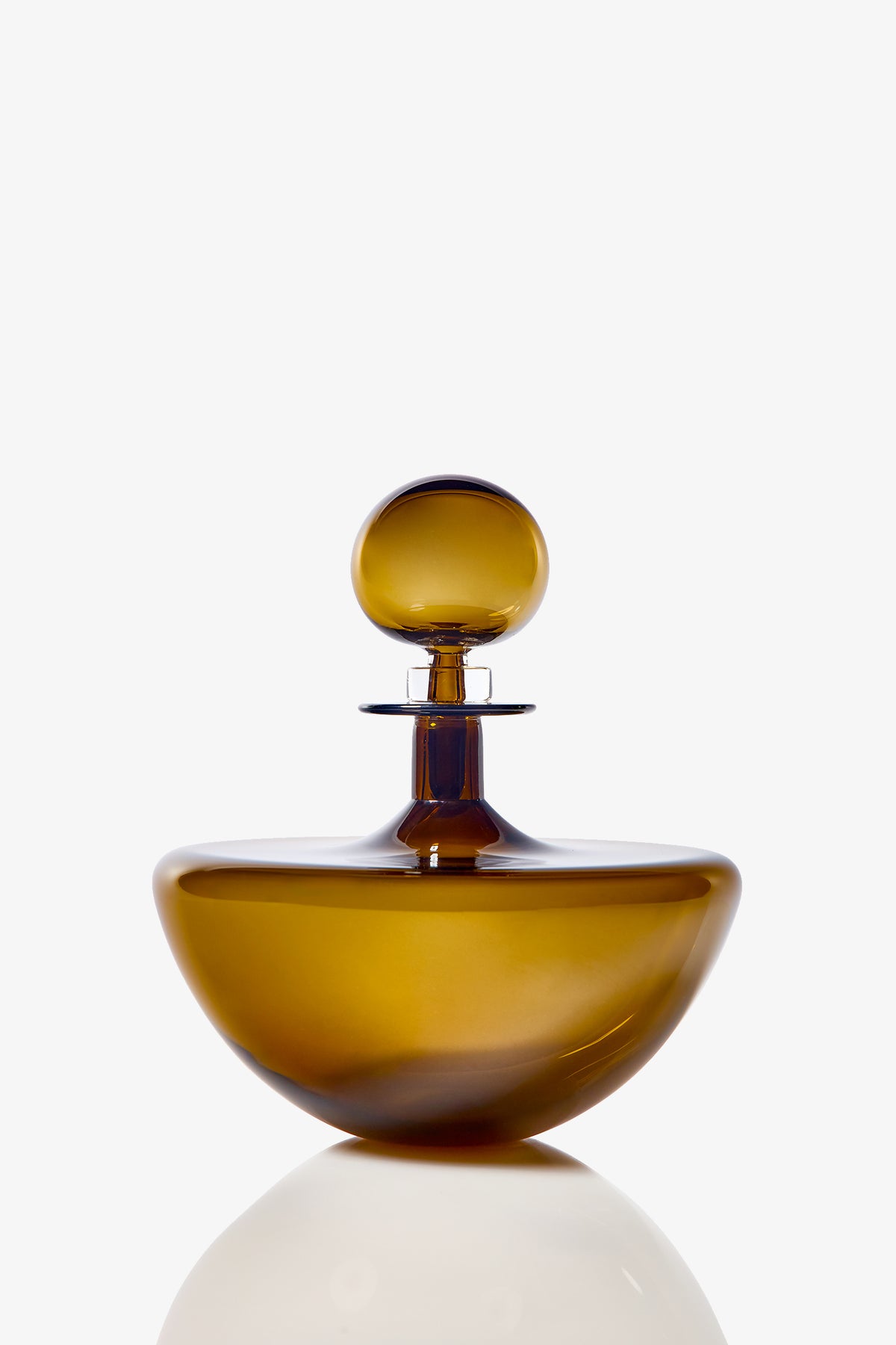 The Small Angelic Bottle, a brown glass perfume bottle with a round stopper and smooth curved design, sits elegantly on a reflective white surface against a plain white background.