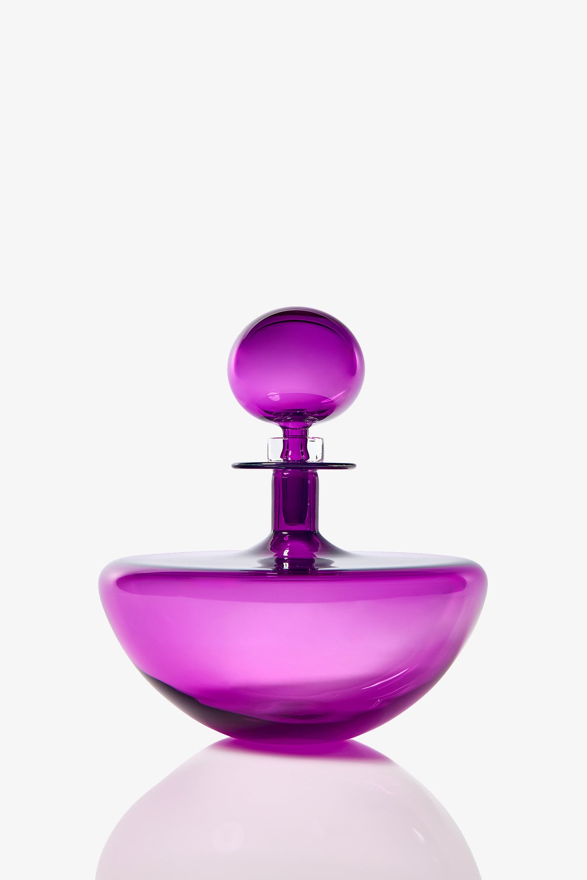 The Small Angelic Bottle, a shiny, translucent purple glass perfume bottle with a round stopper, sits on a reflective surface against a plain white background.