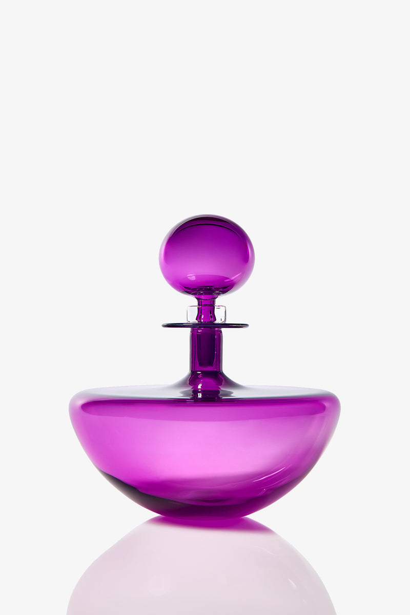 The Small Angelic Bottle, a shiny, translucent purple glass perfume bottle with a round stopper, sits on a reflective surface against a plain white background.