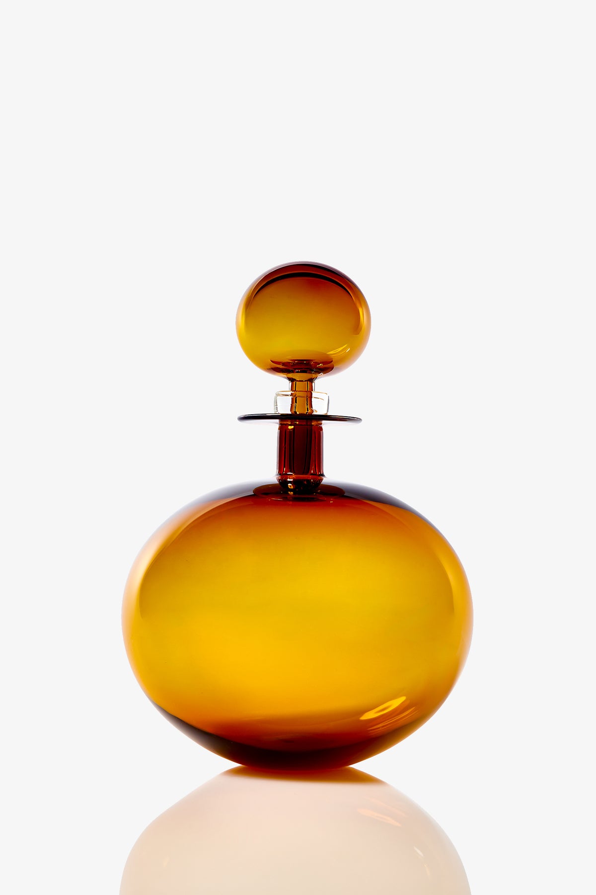 The Small Angelic Bottle, a round amber glass perfume bottle with a matching spherical stopper, rests on a reflective white surface against a plain white background.