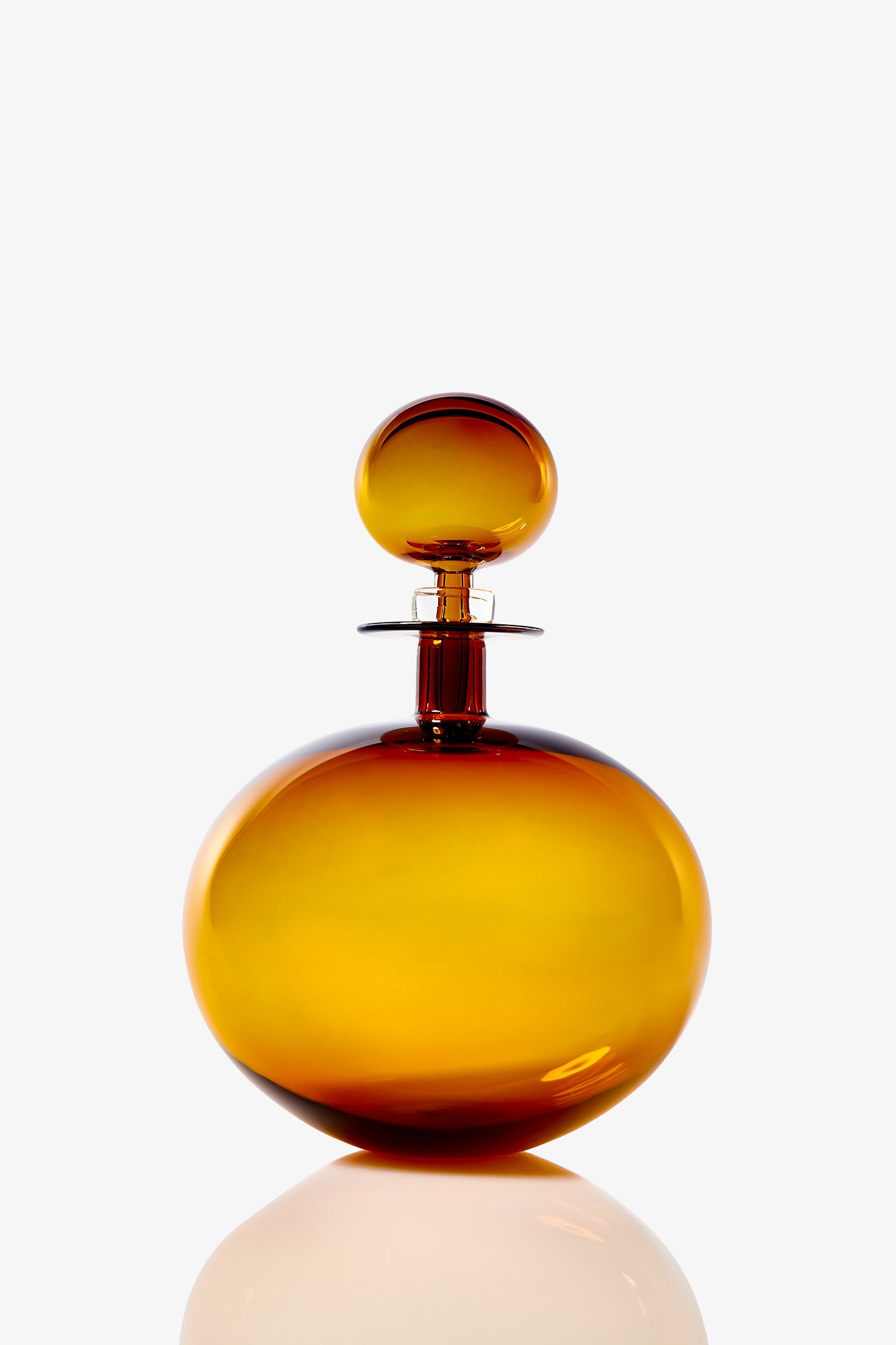 The Small Angelic Bottle, a round amber glass perfume bottle with a matching spherical stopper, rests on a reflective white surface against a plain white background.